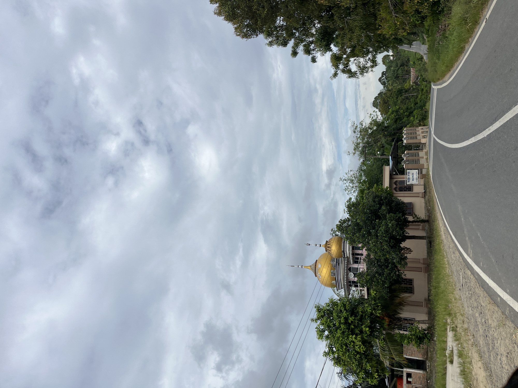 The image shows a mosque in Sukaramai Dua, Indonesia. The mosque has two golden domes and a light-colored building. It is surrounded by trees and vegetation. A paved road with white lines curves in front of the mosque. The sky is overcast with gray clouds. A sign with Arabic writing is visible near the entrance of the mosque. The overall scene depicts a quiet, rural setting.