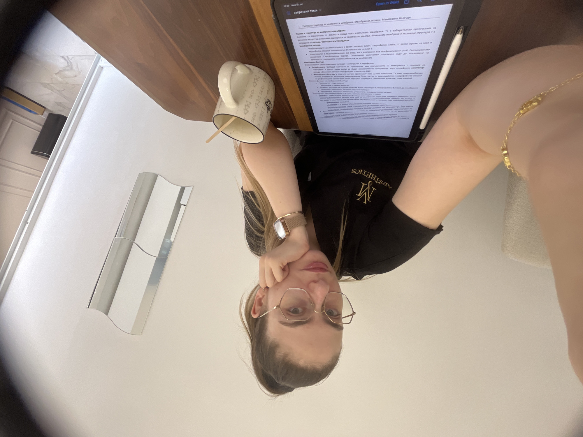 A young woman with a ponytail and glasses is seated at a wooden table, resting her chin on her hand. She is wearing a black t-shirt with a logo. In front of her is a tablet displaying text and a stylus. To her right is a white mug with a swirled pattern and a wooden stirrer. The background shows a white wall, a part of a chair, and a reflective surface. The lighting is bright, suggesting an indoor environment.