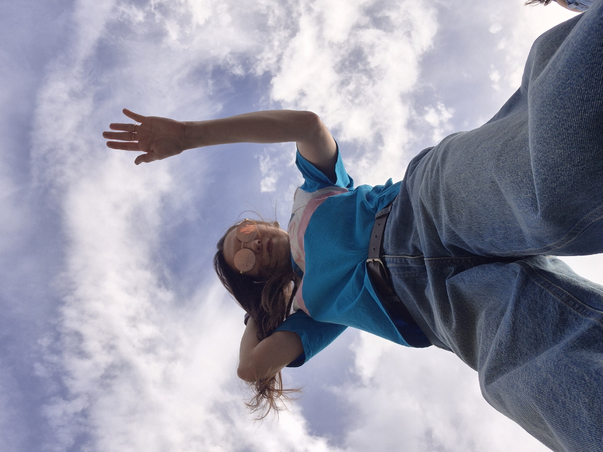 The image is taken from a low angle, looking up at a person against a sky filled with white and light blue clouds. The person is wearing round, rose-tinted sunglasses, a blue t-shirt with a pink stripe, and blue jeans held up by a brown belt. One arm is extended upwards with the hand open, fingers spread, while the other arm is bent and held near the head. The hair is long, brown, and appears to be swept back. The location is not identifiable from the image.