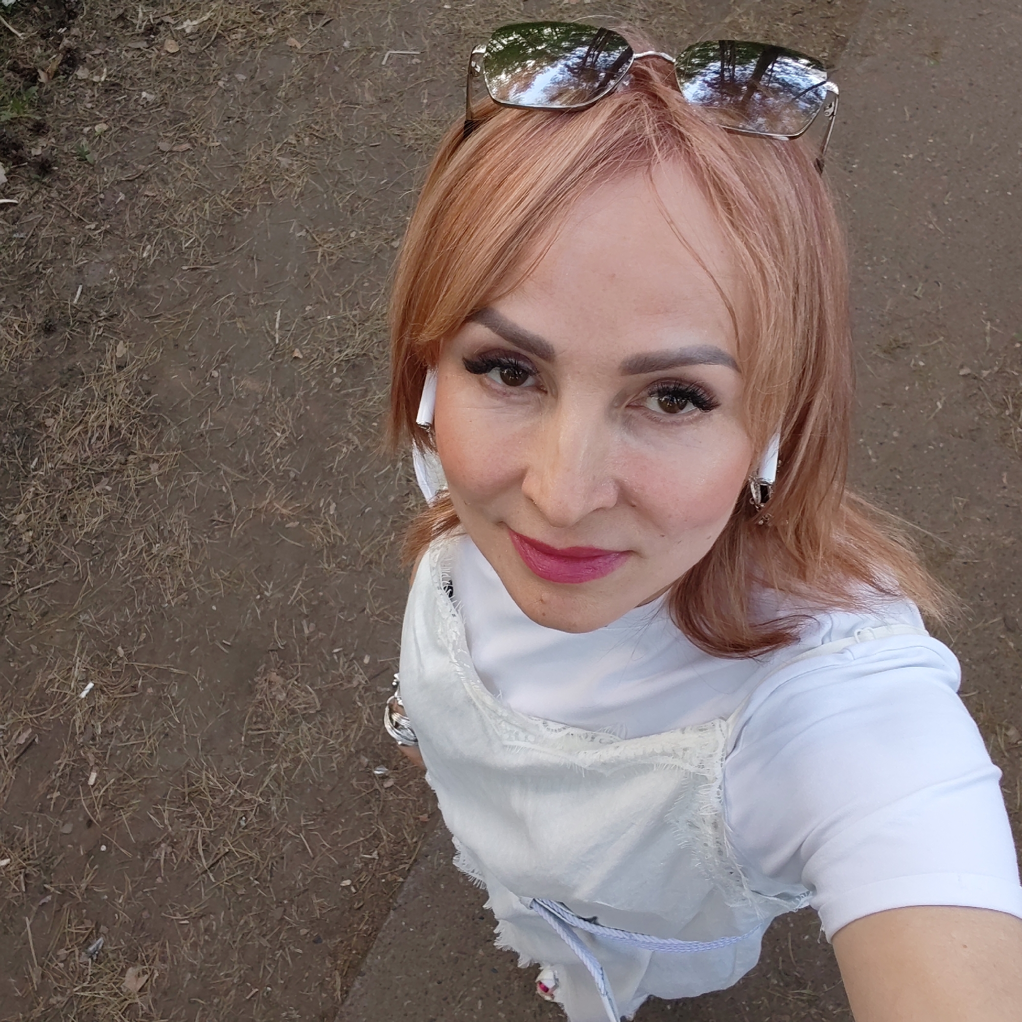 A woman with reddish-blonde hair and sunglasses perched on her head is captured in a selfie taken outdoors. She wears white wireless earbuds and a white top with a layered, textured overlay. The background consists of a ground covered with a mix of dirt and scattered dry grass. The location may be in the city of Chaikovsky, Russia.