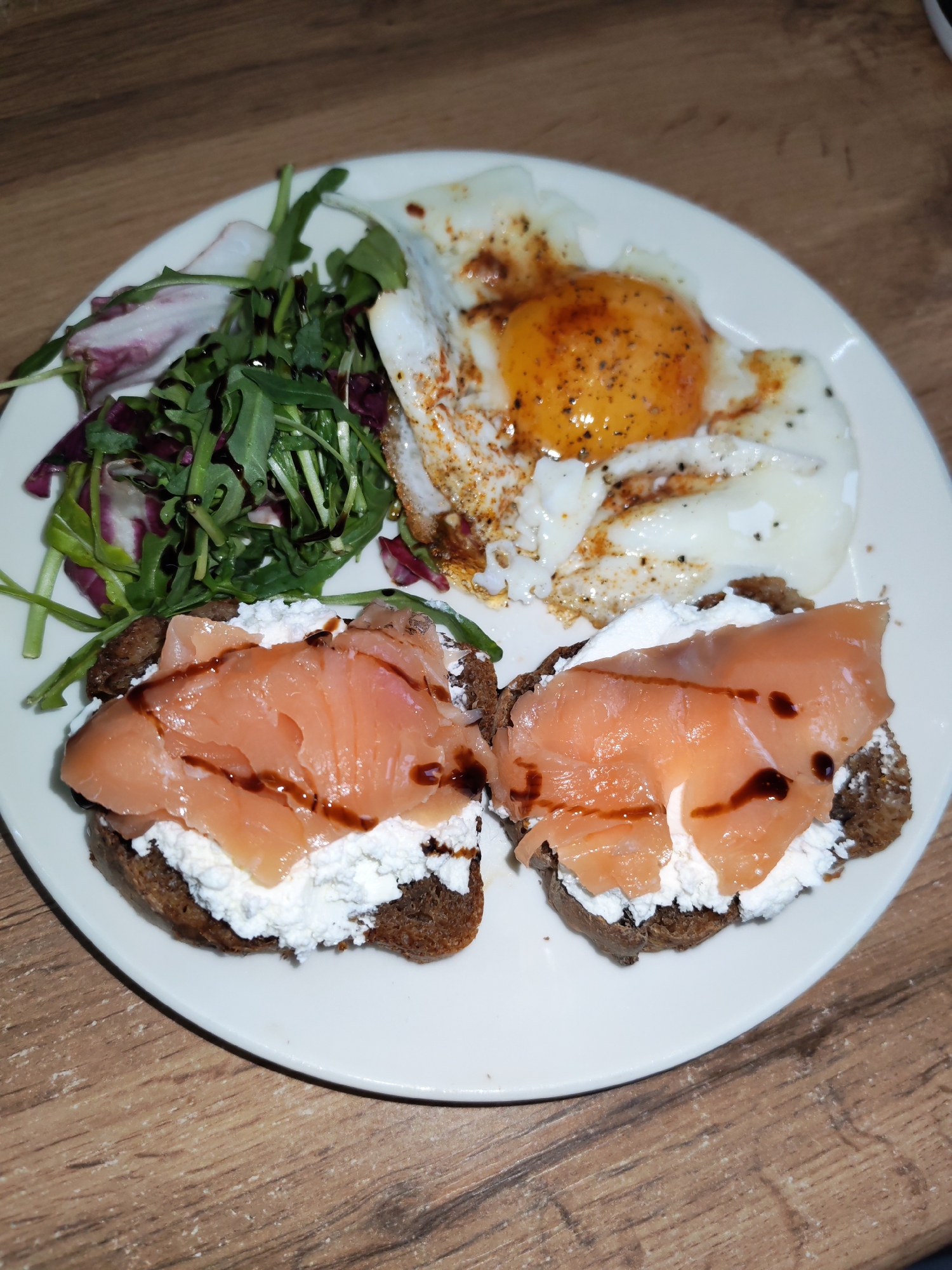 On a white plate, a meal is presented against a wood-grain surface. The meal consists of two open-faced sandwiches featuring dark bread, a layer of white cheese (possibly ricotta or cream cheese), and salmon slices drizzled with a dark sauce. Next to the sandwiches is a small salad of mixed greens and red leaves, also drizzled with the same dark sauce. A fried egg, seasoned with pepper, is positioned on the other side of the plate. No people are visible in the image. The location in Izhevsk, Russia, is unknown from the visual information provided.