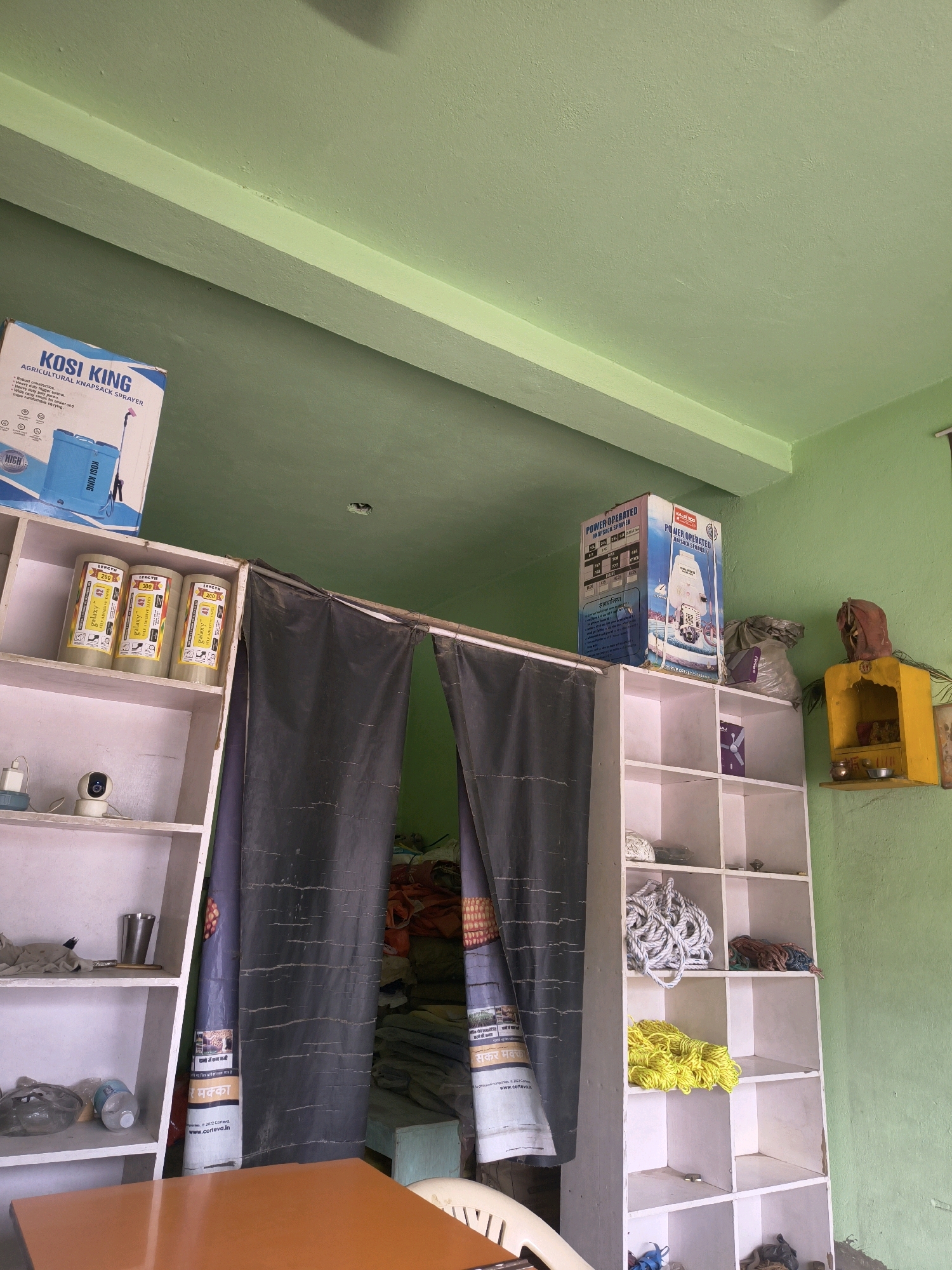 The image shows the interior of a shop located in Purnia, India. The shop appears to be a retail store selling agricultural supplies and equipment. The walls are painted a light green, and the ceiling is also green, with a white beam running across it.
On the left side of the image, a blue and white box labeled "KOSI KING AGRICULTURAL KNAPSACK SPRAYER" is visible. Below it, a white shelf unit displays several large cylindrical containers, possibly for pesticides or fertilizers, with labels that include "GALAXY". A small security camera is also on this shelf.
In the center, a dark curtain hangs, partially obscuring items behind it. A tall, white shelving unit stands to the right of the curtain. This unit holds various items, including coils of rope (white and yellow) and a small bag of what appears to be soil or fertilizer. A small yellow shrine with deities is mounted on the wall above the rightmost shelf.
In the foreground, the corner of an orange table is visible, and a white chair is partially seen. The overall impression is of a well-stocked shop, with goods neatly arranged on shelves.