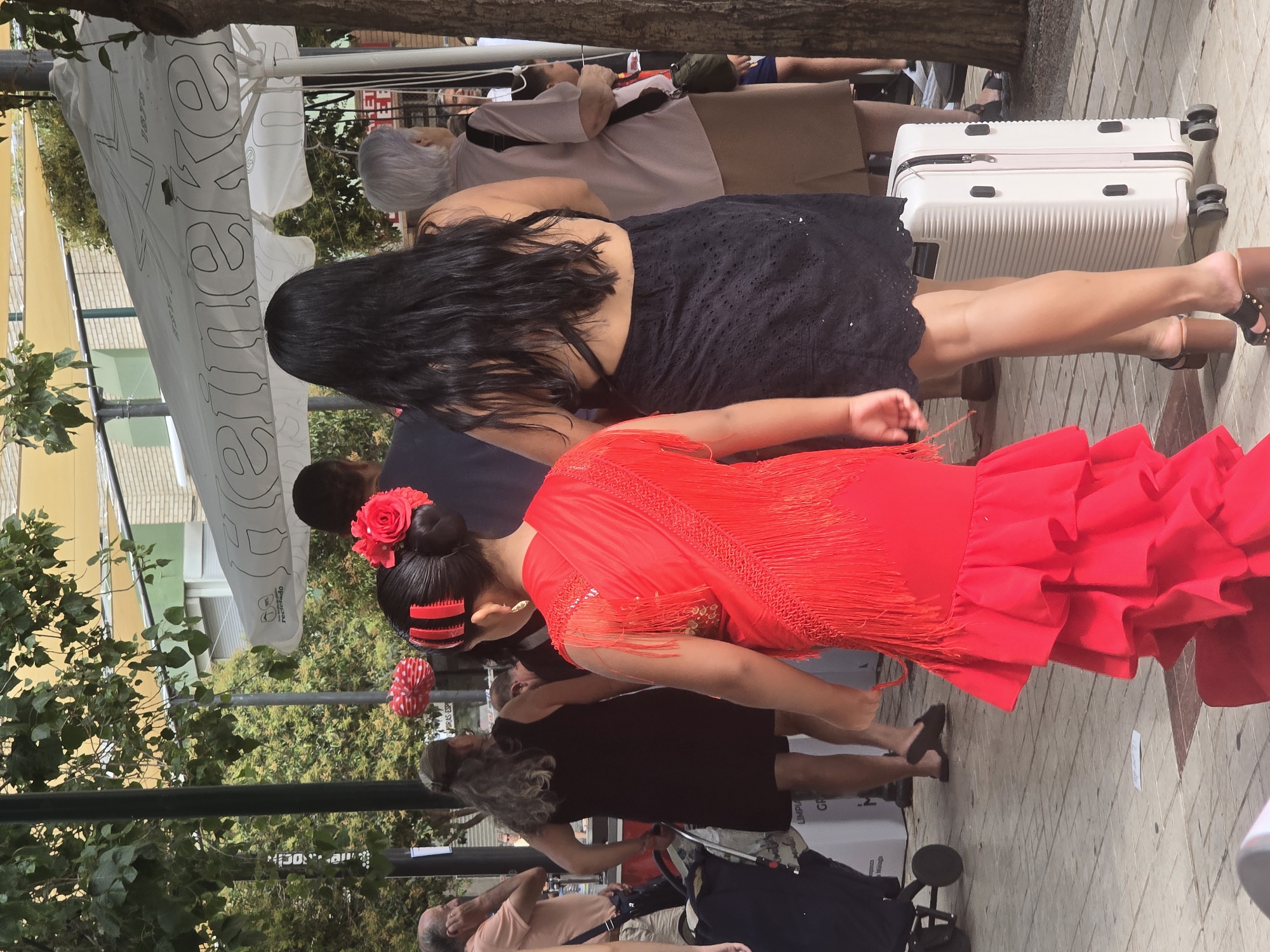 The image captures a lively street scene in Málaga, Spain, likely during a festive event, with a focus on a young girl in a vibrant red flamenco dress. She is positioned in the foreground, her back to the camera, showcasing the intricate layers of her ruffled skirt and fringed shawl. Her hair is styled with a red comb and a prominent red rose. Behind her, another woman in a black, open-back dress walks with a white suitcase on wheels. In the background, a diverse crowd of people are visible, some shaded by large white umbrellas bearing the Heineken logo. The ground is paved with interlocking stones. Trees and buildings, including what appears to be a green-tiled structure, are present in the background. The overall atmosphere suggests a celebratory and possibly crowded public gathering.