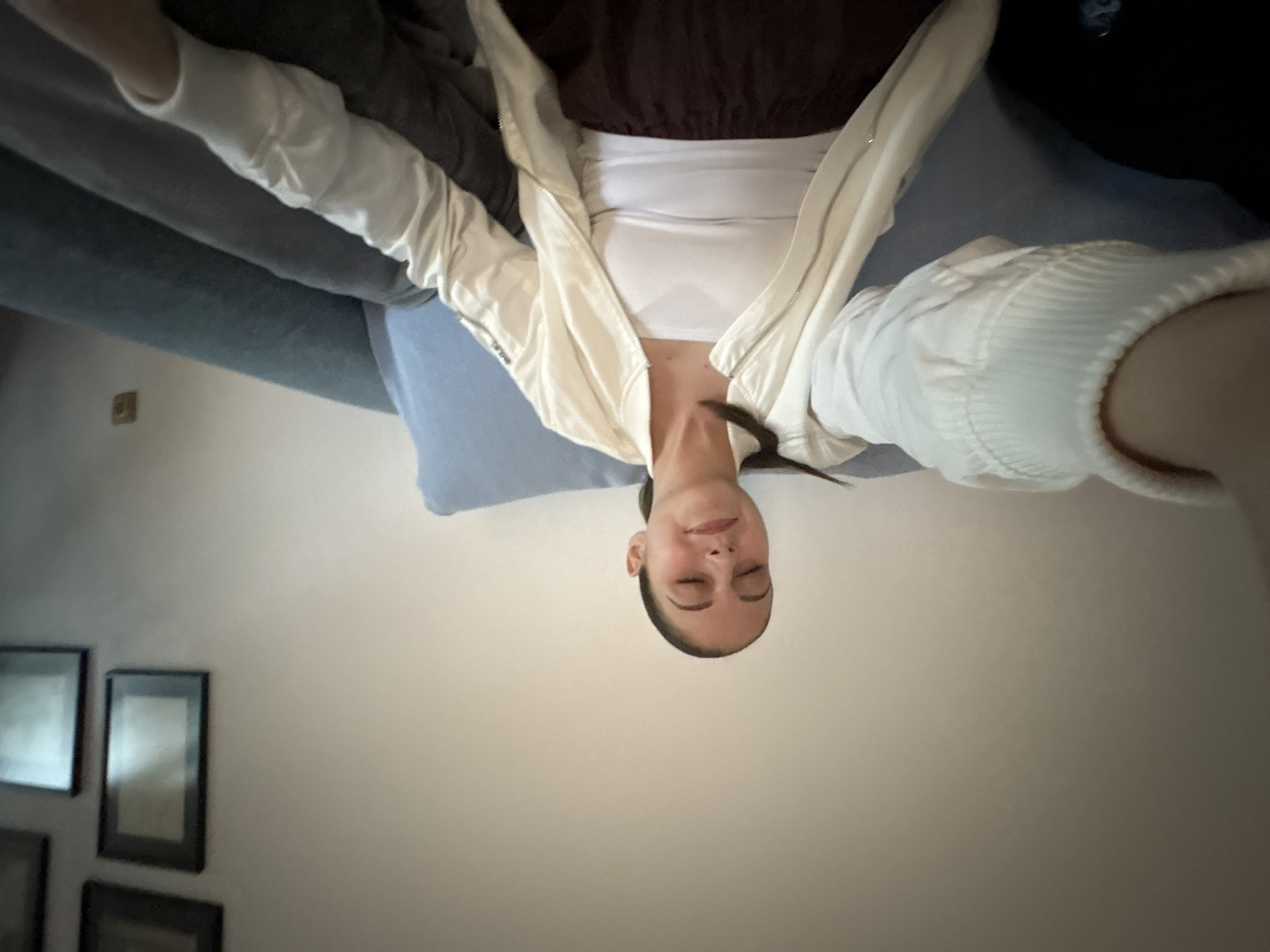 A person is lying on a blue sofa with their head toward the bottom of the image, facing upward. They are wearing a white tank top, a brown top, and a white jacket. Their eyes are closed. The background includes a beige wall with three framed pictures. The location is not discernable as Burgas, Bulgaria.