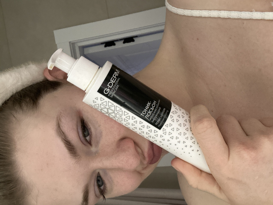A woman holds a white bottle of G-Derm Professional Skincare Tonic Lotion. The bottle is white with a black section bearing the brand name and product information in Russian and English. The text includes "ТОНИК-ЛОСЬОН," "Гипосенсибилизирующий," and "TONIC-LOTION Hypersensitizing." A pattern of light-colored triangles adorns the lower section of the bottle. The woman has a white headband and is wearing a white strap top.