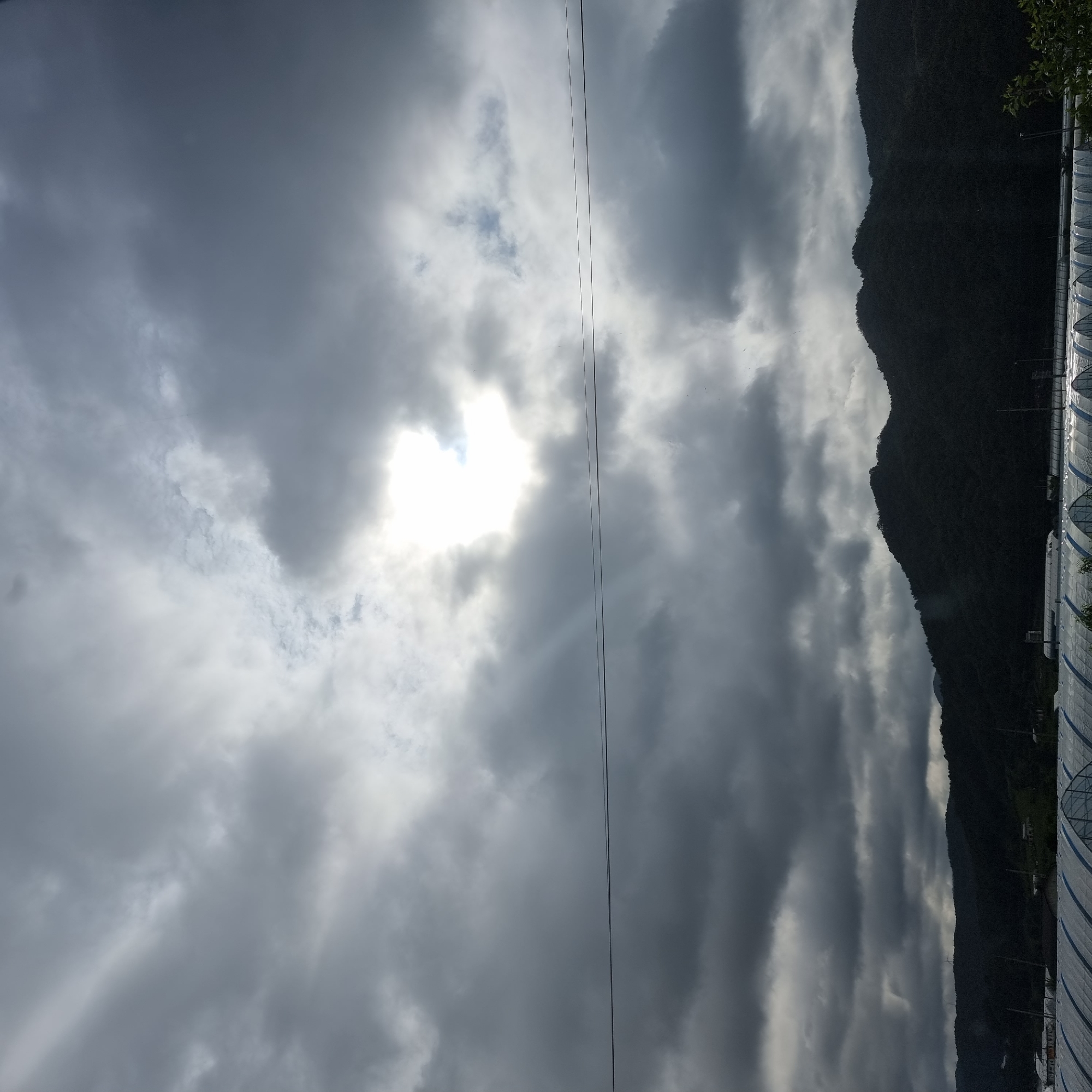 The image depicts a cloudy sky over a rural landscape. The sun is partially visible through a break in the clouds, casting rays of light. In the foreground, there are several greenhouses with white coverings, suggesting agricultural activity. A power line stretches horizontally across the middle of the frame. In the background, dark, forested hills rise against the sky. The location is identified as Dunnae-myeon, South Korea. There are no people or specific actions captured in the scene, which is a static environmental shot.