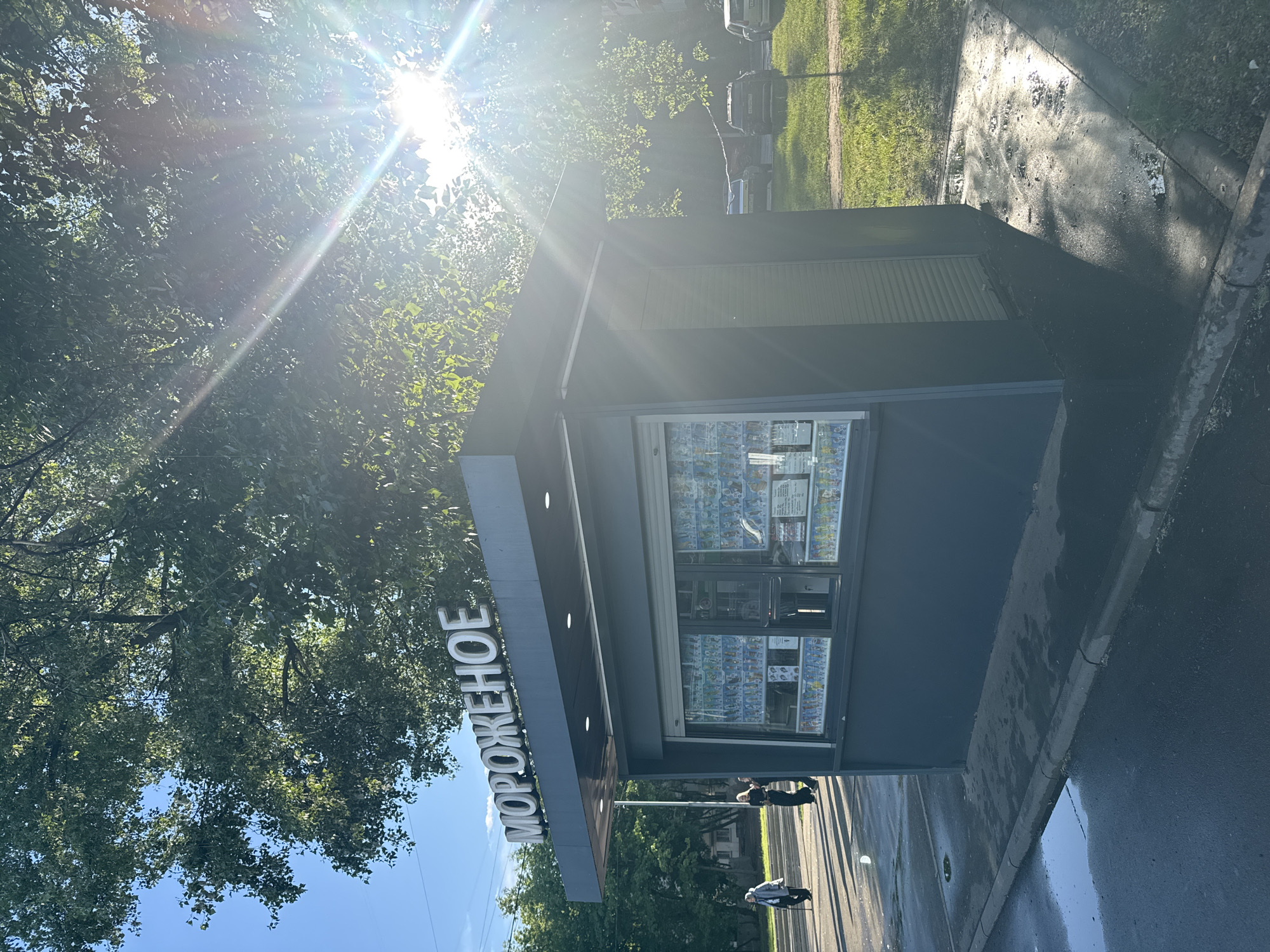 The image shows an ice cream kiosk, with "MOROZHENOYE" (ice cream in Russian) spelled out in large letters on the awning. The kiosk is a modern, box-like structure with a glass display case full of frozen treats. Two people are walking in front of the kiosk. The sun is bright, causing a strong glare through the trees. The location appears to be in a city, likely Moscow, Russia. The street looks wet, possibly due to rain. Cars are parked in the background amidst green vegetation.
