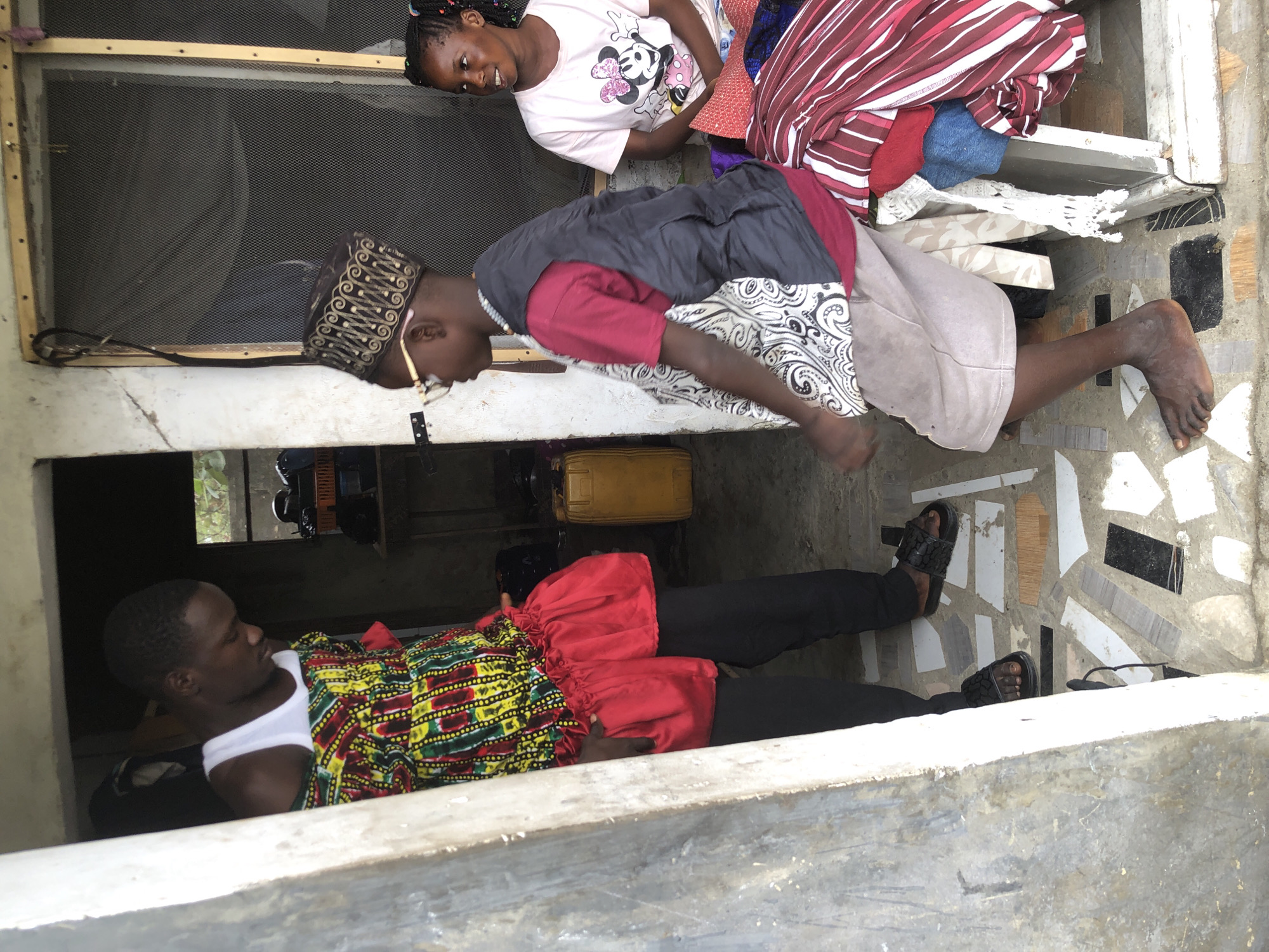 In Ado, Nigeria, a man wearing a white tank top and a vibrantly patterned shirt with a red ruffled hem stands in a doorway. He is looking down and to his left. To his right, a young boy wearing glasses and a decorative patterned cap, a dark vest over a red t-shirt, and light-colored shorts, is looking to his left. Behind the boy, a young woman in a pink t-shirt with a Minnie Mouse graphic smiles while looking to her right. The floor is decorated with a mosaic of colorful and varied tile fragments. Several items of clothing, including striped and red fabrics, are piled to the right of the woman. A yellow plastic barrel is visible in the background, along with other indistinct objects and structures. The overall scene appears to be an outdoor or semi-enclosed living space.