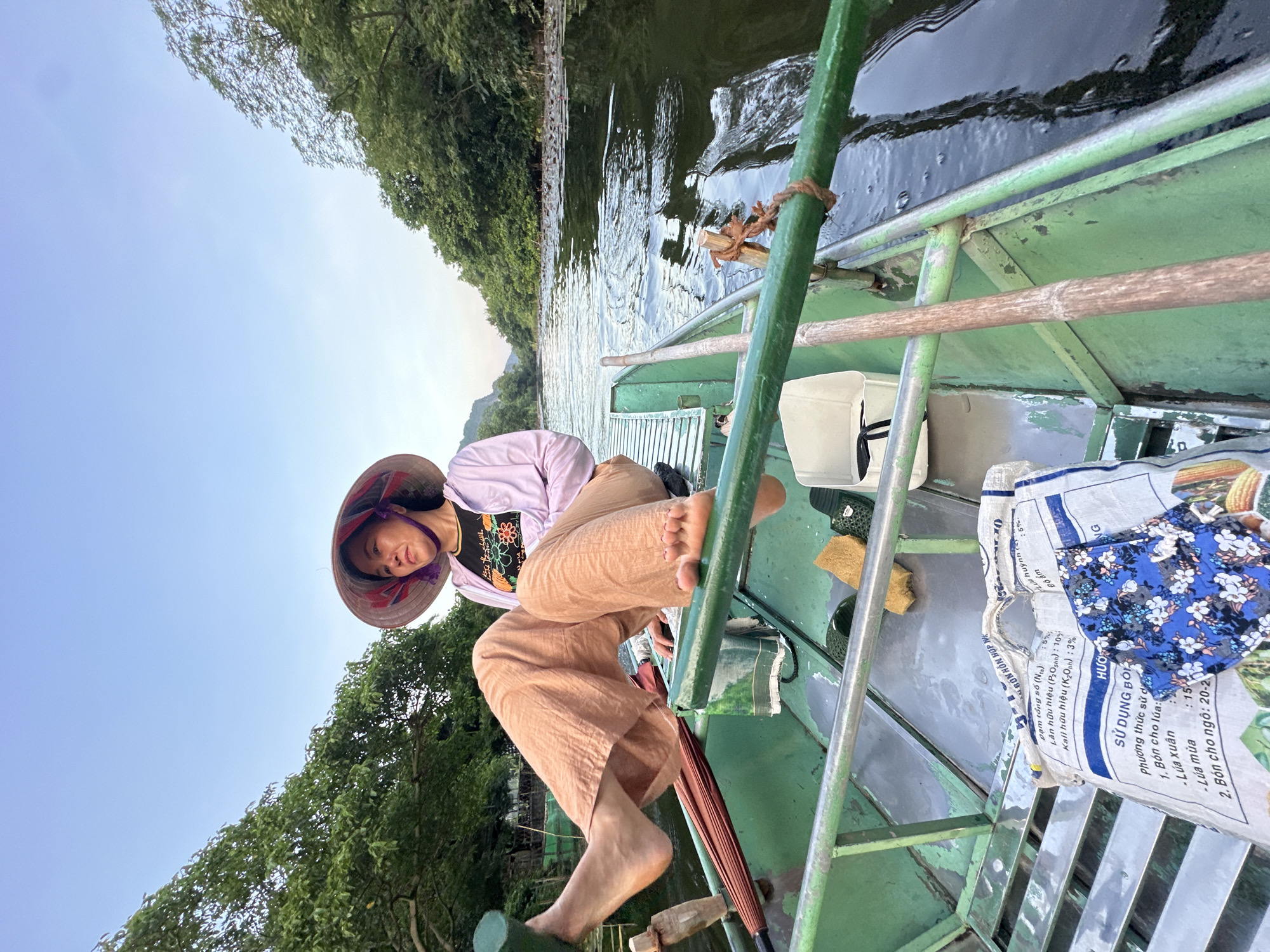 A woman wearing a conical hat and a light purple jacket is seated on a green wooden boat on a river. She is looking to her left with a relaxed expression. Her legs are crossed, with her bare feet visible. The boat is filled with various items, including bags of fertilizer, an umbrella, and a white bucket. The river is surrounded by lush green trees and vegetation. The sky is a pale blue with some clouds. The scene appears to be in a natural setting, possibly a tourist area in Vietnam, given the conical hat and the style of the boat. The location is identified as Nam Hoa Lư Ward, Vietnam.