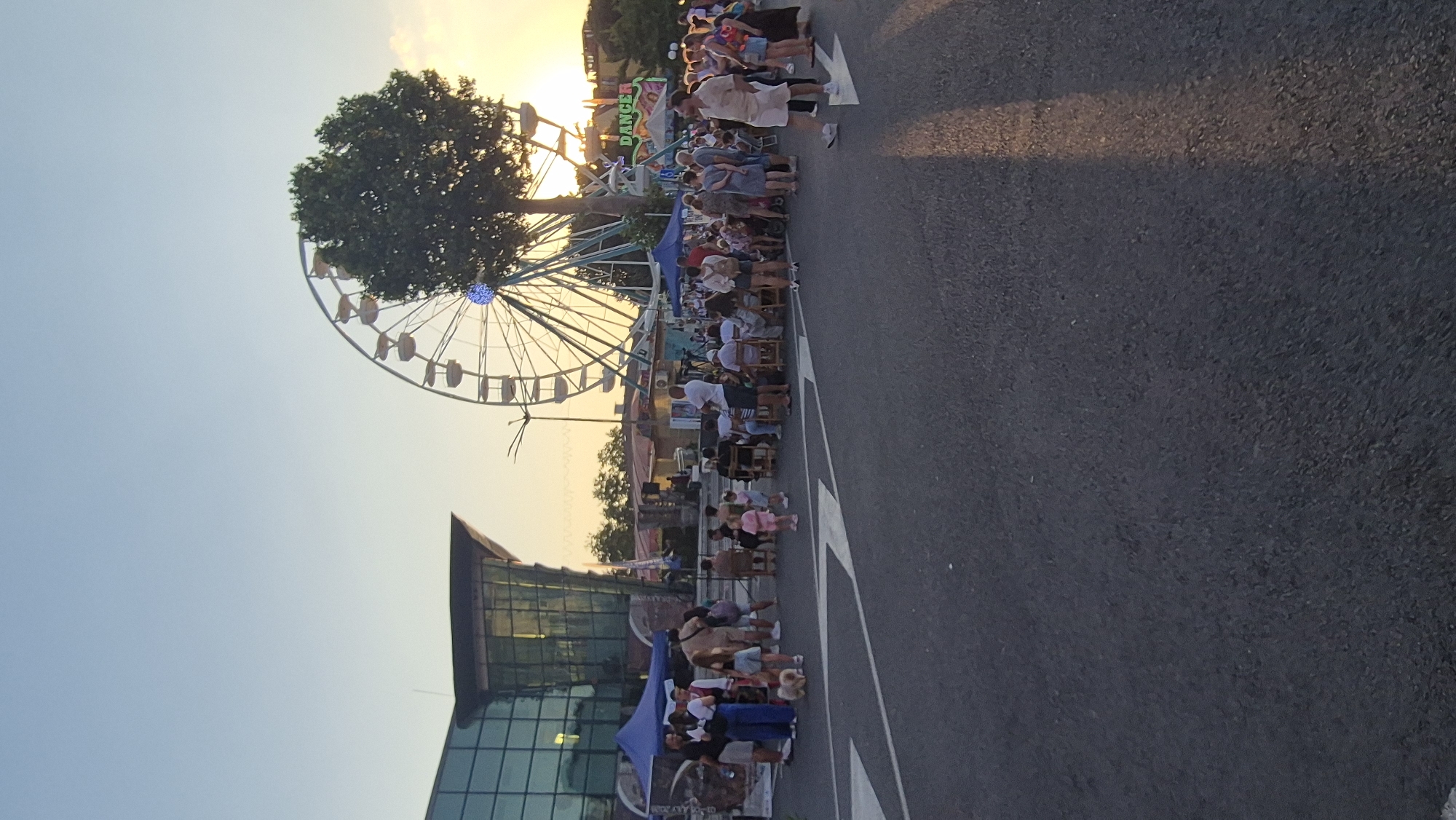 This image captures an outdoor event near the "DANSKY" amusement ride in Pomorie, Bulgaria, likely at dusk. A Ferris wheel is prominently featured in the background, partially obscured by a large tree. Several people are gathered, some sitting on chairs, others standing, suggesting a festive atmosphere. A modern building with glass windows is visible to the left, possibly a shopping center or public facility. Tents are set up, indicating stalls or activity areas. The ground is paved asphalt, with white lane markings visible in the foreground. The sky is a soft blue transitioning to warm, orange hues from the setting sun.