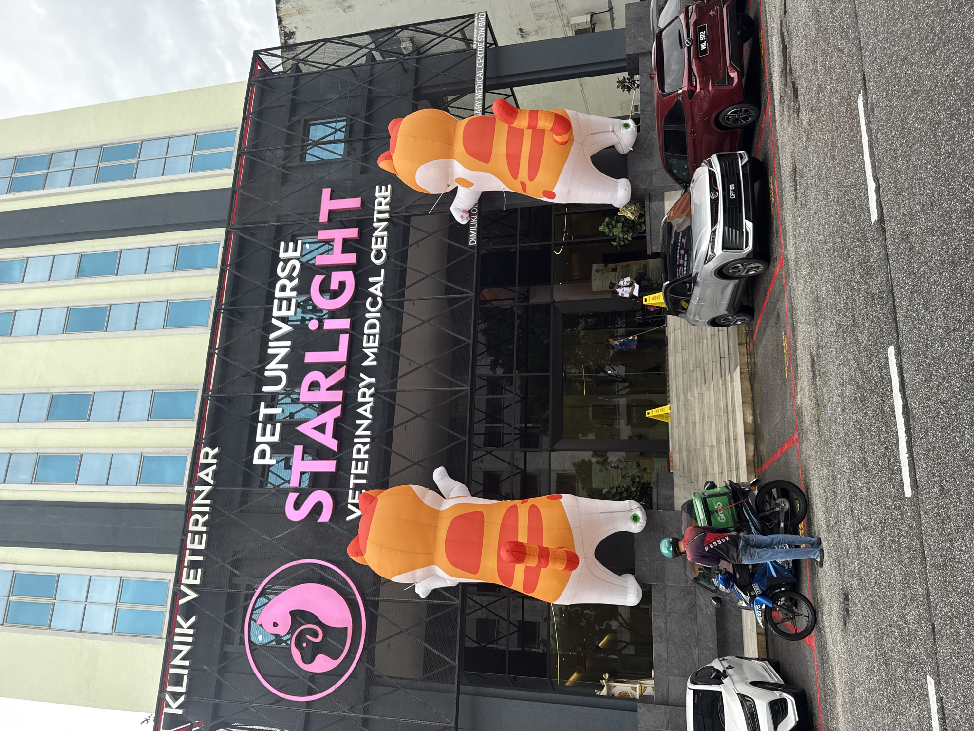 The image shows the front of a veterinary medical center called "Pet Universe Starlight Veterinary Medical Centre," located in Ampang Jaya Municipal Council, Malaysia. The building has a modern black facade with large inflatable cat decorations attached to the exterior. A sign displays the clinic's name and logo, with a pink circle containing a silhouette of a cat and a dog.
Several vehicles are parked in front of the building, including a red car, a white car, and a silver car. A person on a motorcycle with a Grab delivery bag is visible near the entrance. The overall environment appears to be an urban street with clear weather.