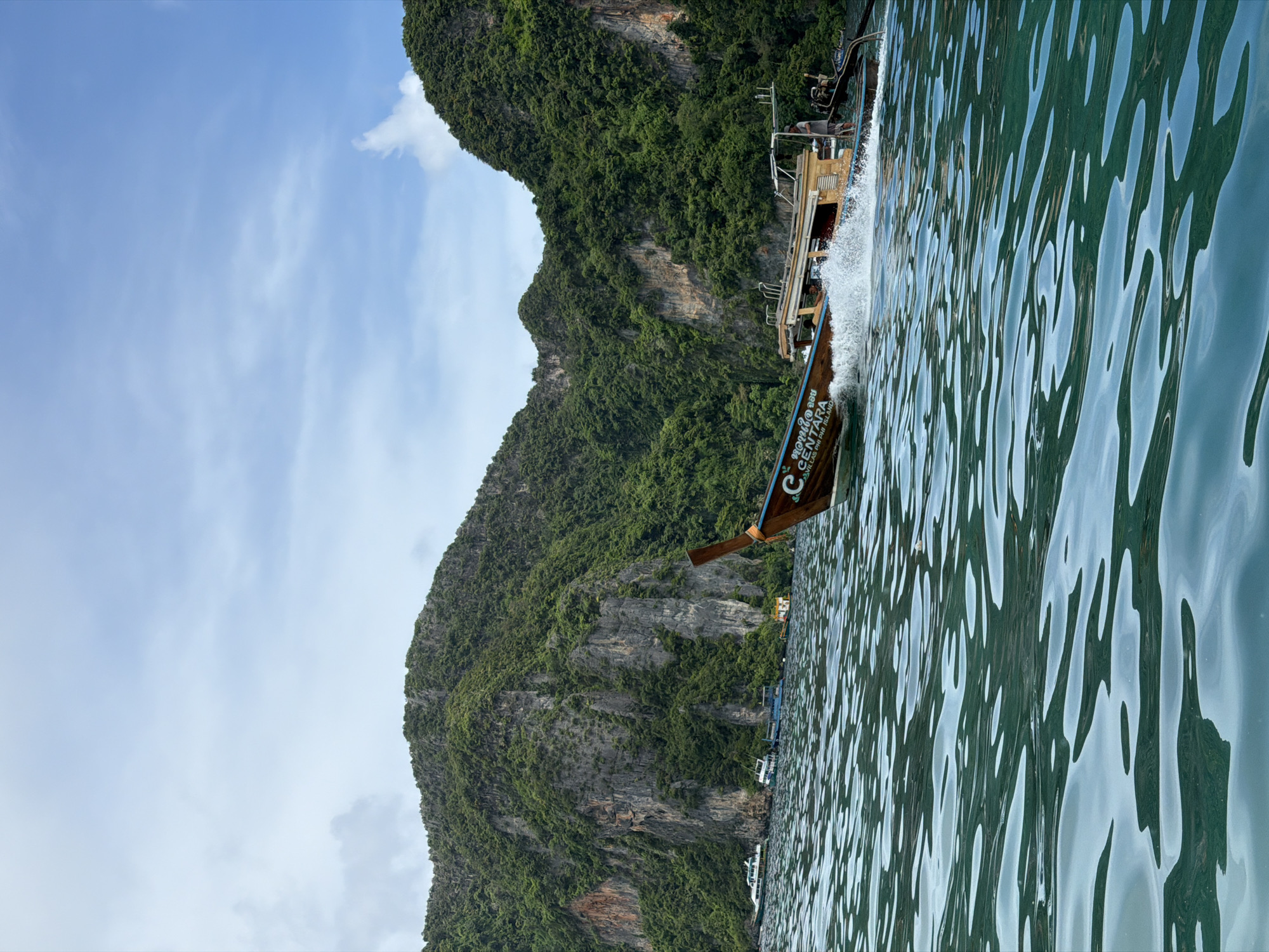 In Ao Nang, Thailand, a longtail boat speeds across turquoise water, leaving a white wake. The boat, adorned with the word "CENTARA," is wooden with an extended bow and stern. Several other boats, including a larger white vessel, are anchored or moving slowly in the distance. Towering, lush green limestone cliffs rise dramatically from the water behind the boats, their rocky faces partially obscured by dense vegetation. The sky above is a mix of blue with scattered white clouds.