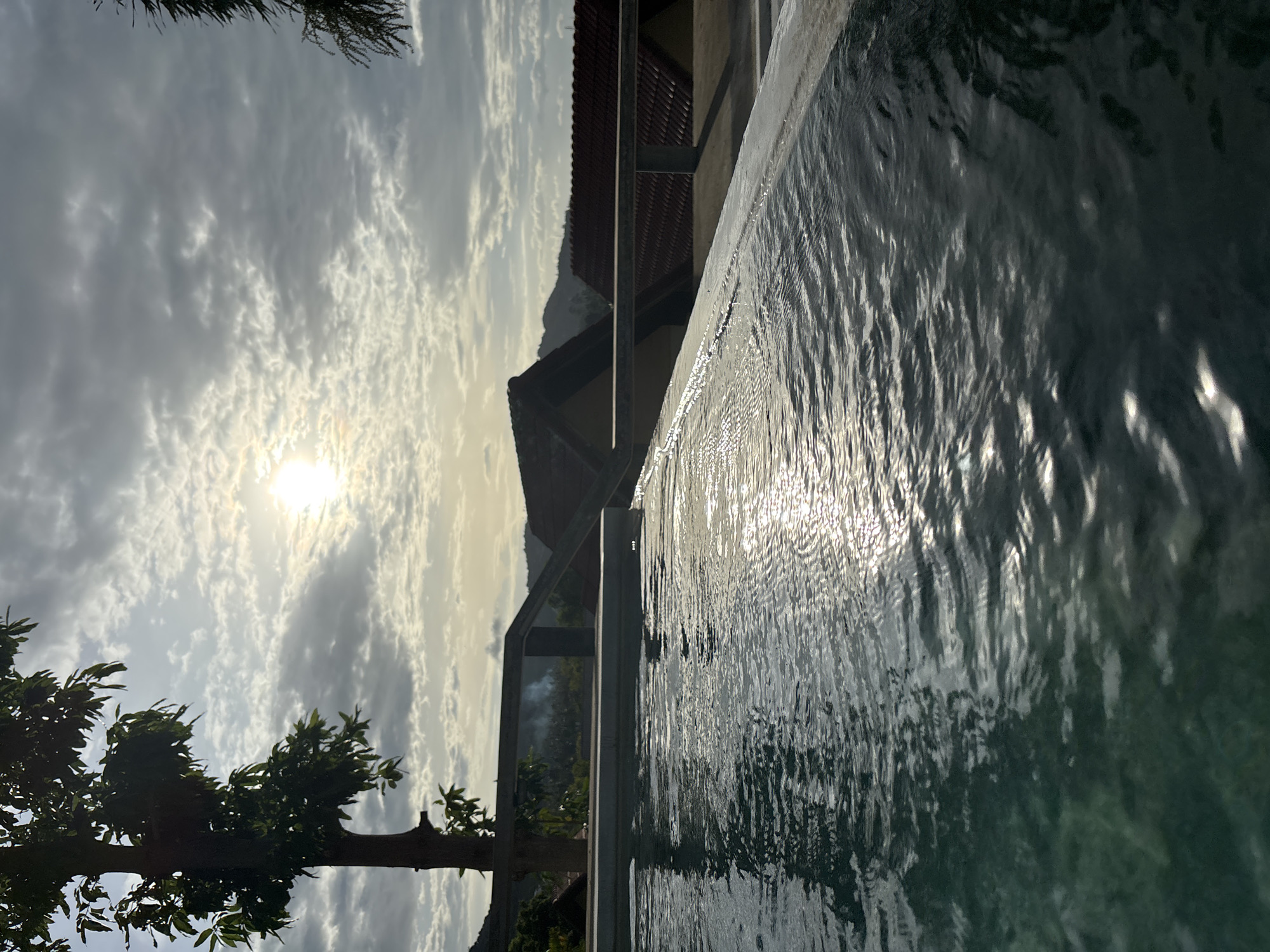 A low-angle shot captures the edge of a swimming pool with rippling water in the foreground, reflecting the sunlight. Beyond the pool, a tree with dark leaves stands on the left, silhouetted against a cloudy sky. In the background, several buildings with reddish-brown tiled roofs are visible, nestled amongst hills. The scene is set at dawn or dusk, with soft, diffused light illuminating the sky and casting gentle reflections on the water. The location is identified as Baan Bang Rak, Thailand.