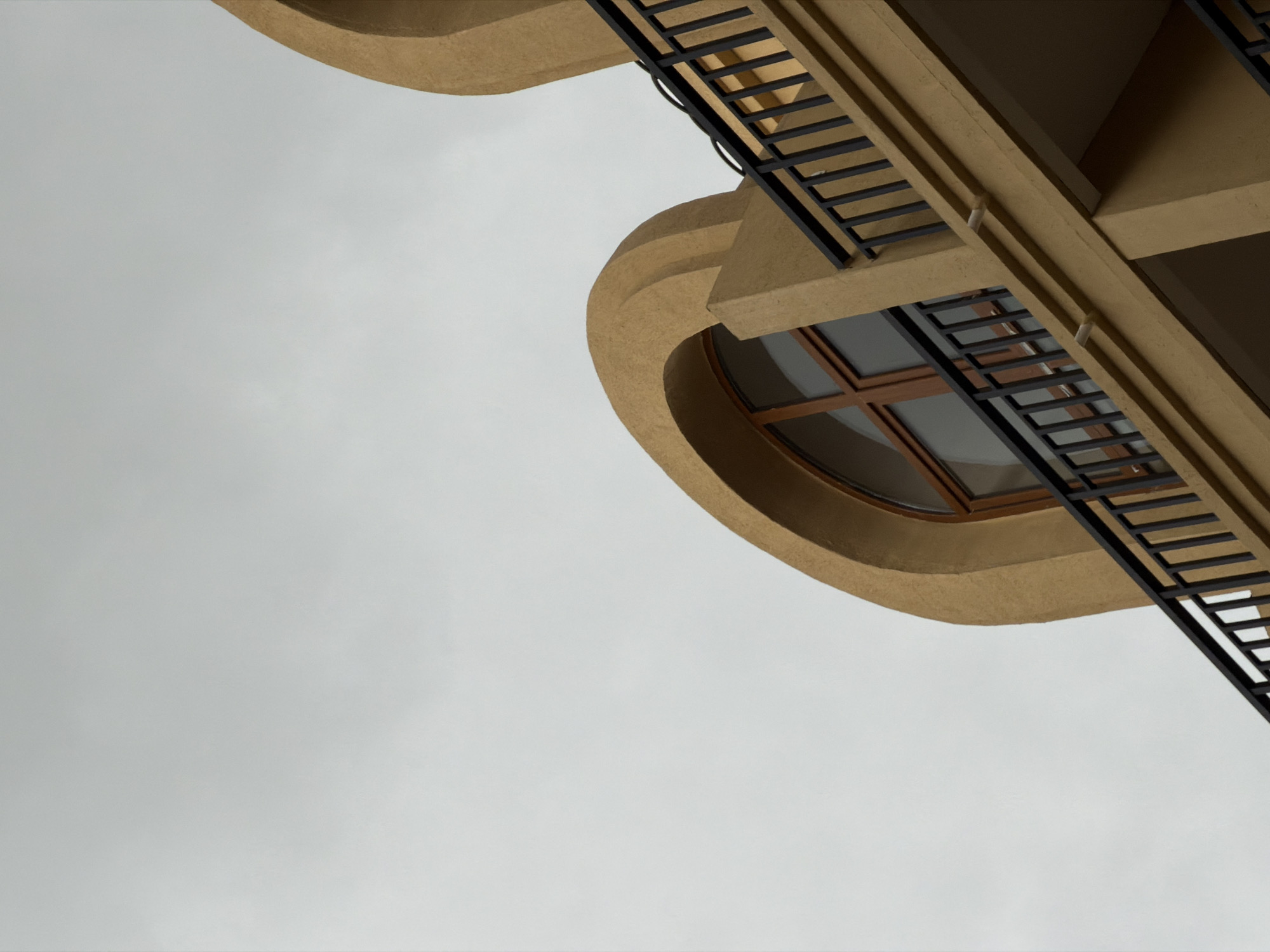 A low-angle shot reveals a section of building in Tbilisi, Georgia, against a grey sky. The building features tan-colored, curved architectural elements and a distinctive oval window with a brown frame. A black metal fire escape with horizontal rungs ascends along the building's facade. The perspective of the shot emphasizes the upward angle and the architectural details of the structure.