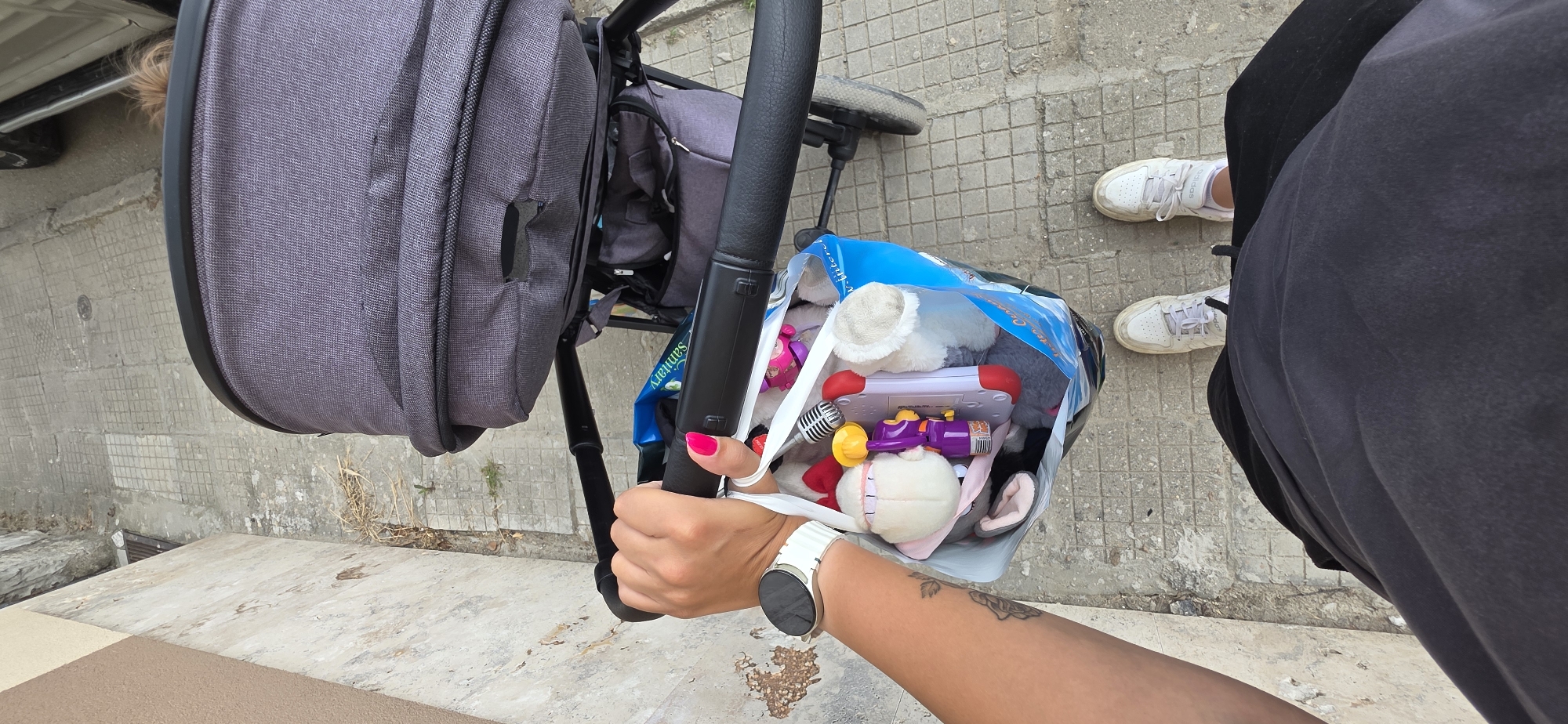 The image provides a high-angle, first-person view of a person standing outdoors next to a baby stroller, holding a bag of toys. The setting is a paved area in Plovdiv, Bulgaria, characterized by grey concrete blocks, with some dirt and dried grass visible on the ground.
The subject is partially visible, wearing dark pants and white Adidas sneakers. Their right arm and hand are prominent, showing a white smartwatch on the wrist and bright pink nail polish. A floral tattoo is also visible on the forearm. This hand is grasping the black handle of a grey baby stroller.
Attached to the stroller or held by the person is a clear plastic bag filled with numerous colorful toys. These include various plush toys, a red and white electronic toy, a purple and yellow toy, and a silver toy microphone. Visible text on the bag includes "Sammary," "Winter," "Inter," and "Quality."
The scene suggests an everyday activity, likely an outing with a child, where toys are being transported. The bright, natural light indicates it is daytime with clear or partly cloudy weather. A car bumper is faintly visible in the upper left, hinting at a street or parking area nearby.