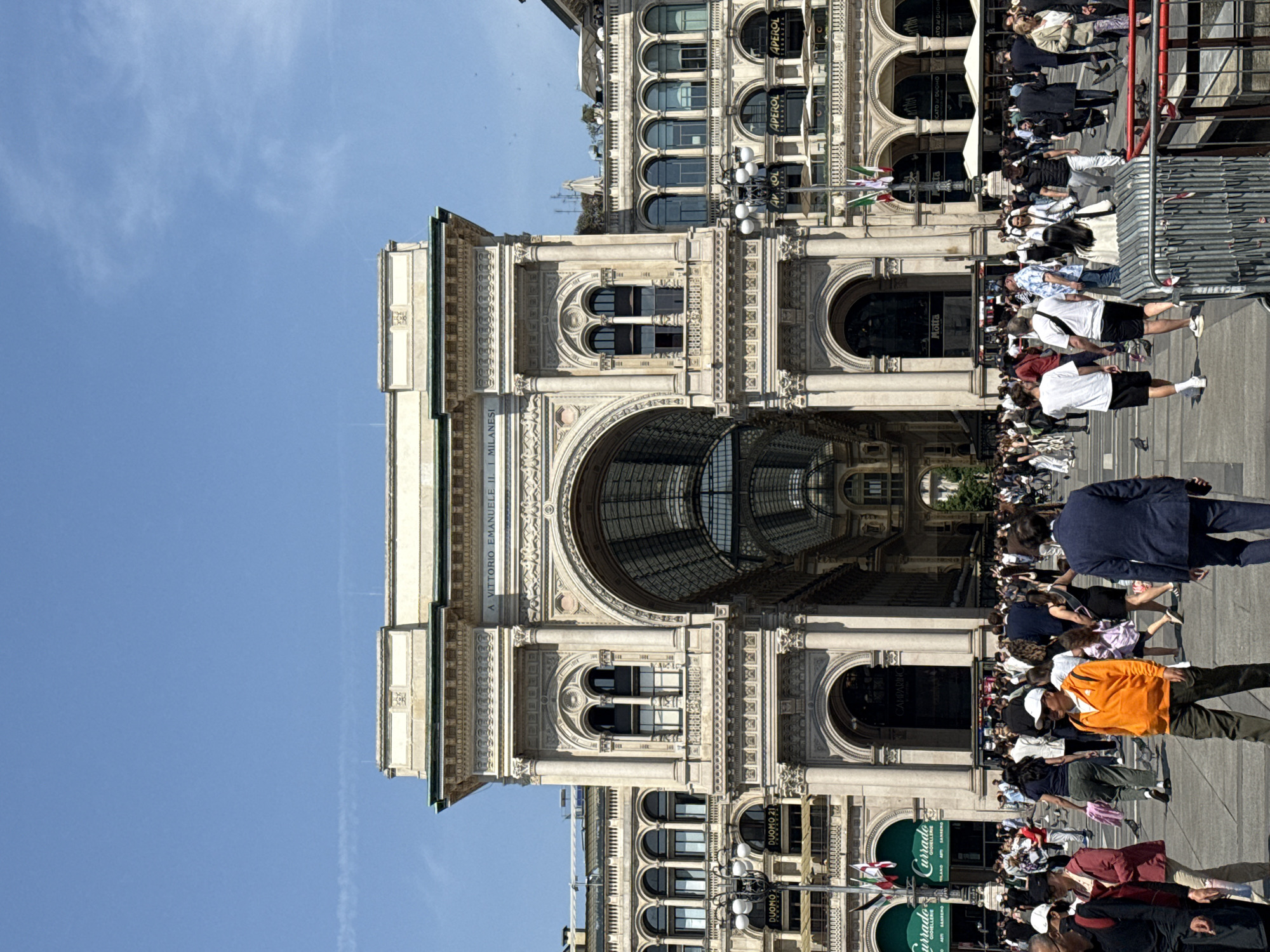 The image captures a bustling daytime scene at the iconic Galleria Vittorio Emanuele II in Milan, Italy. The grand, ornate facade of the 19th-century gallery dominates the left side of the frame, showcasing its classical architecture with elaborate arched windows, intricate carvings, and decorative pilasters. The central archway prominently features the inscription "A VITTORIO EMANUELE II" and offers a glimpse of the gallery's impressive glass and iron ceiling within.
Numerous pedestrians fill the foreground and midground, walking and congregating on the paved area outside the gallery, suggesting a lively atmosphere typical of a popular tourist destination. The weather is bright and sunny, indicated by a clear, vibrant blue sky that occupies a significant portion of the background.
Visible details include Italian flags hanging from poles near the building, and various commercial signs for establishments such as "APEROL TERRAZZA," "DUOMO 21," "Motta," "CAMPARI," and "Currado Gioiellerie" (with "Currado SANREMO" also visible). Partial text like "MILANESI" and "ICATO DUOMO" can also be discerned. Metal barriers or stanchions are present on the right side of the frame, possibly for crowd control or an event. The overall impression is one of a vibrant, sunny day at a significant architectural and commercial landmark.