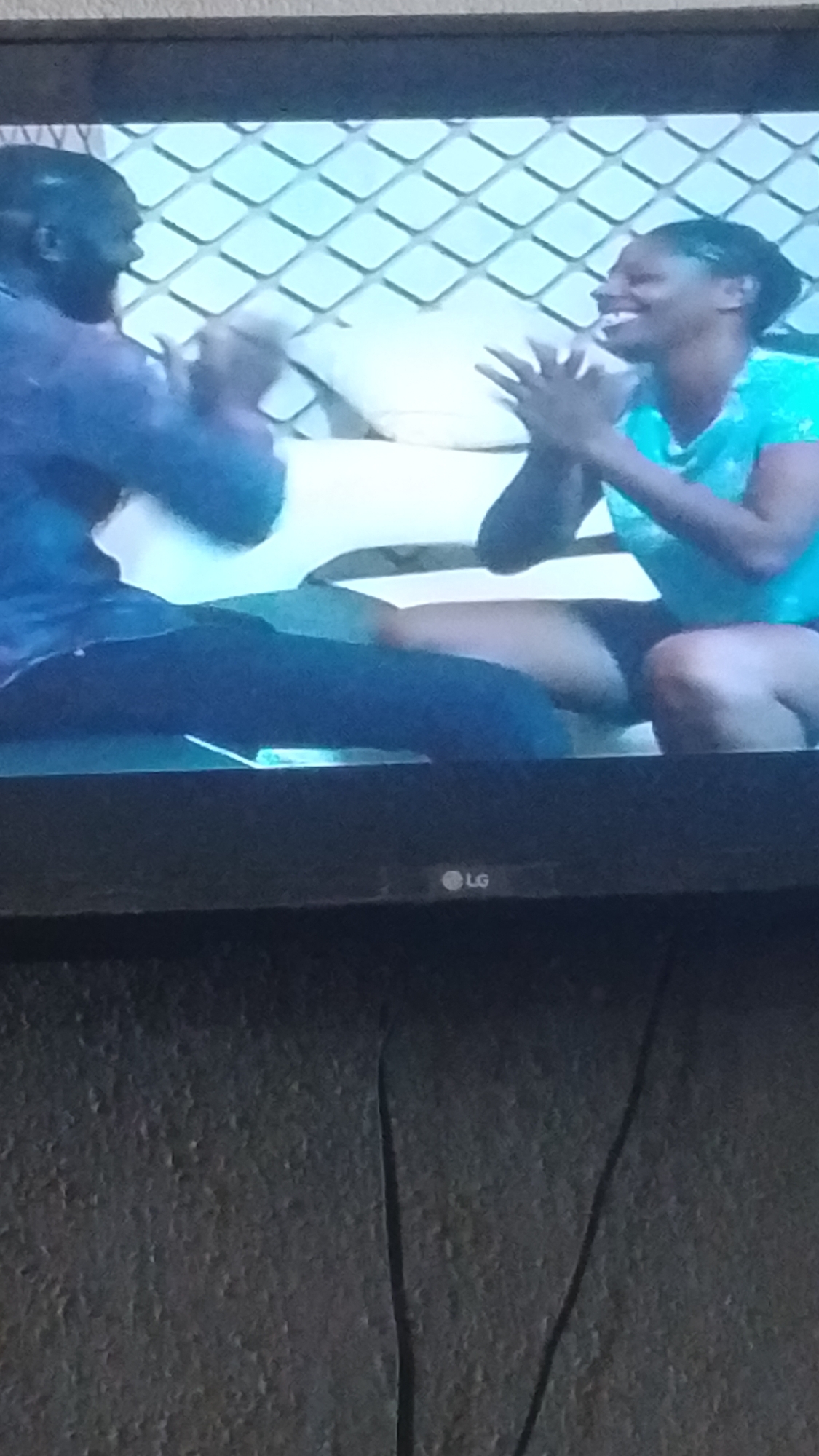 The image captures a moment on what appears to be a television screen, showing two individuals, a man and a woman, engaged in an interaction. The man, positioned on the left, is wearing a dark blue shirt and appears to be leaning forward. The woman, on the right, is wearing a teal-colored top and is smiling. Both individuals have their hands raised, suggesting a playful or interactive gesture like clapping. They are seated on a couch or similar furniture, with a white pillow visible between them. The background features a repeating diamond pattern in a light blue hue, possibly indicating a decorative wall or screen. The lower portion of the television screen shows an LG logo. The scene is likely indoors, given the controlled lighting and the presence of a television. There is no visible indication of the specific location within Lokoja, Nigeria, beyond the context provided in the prompt.