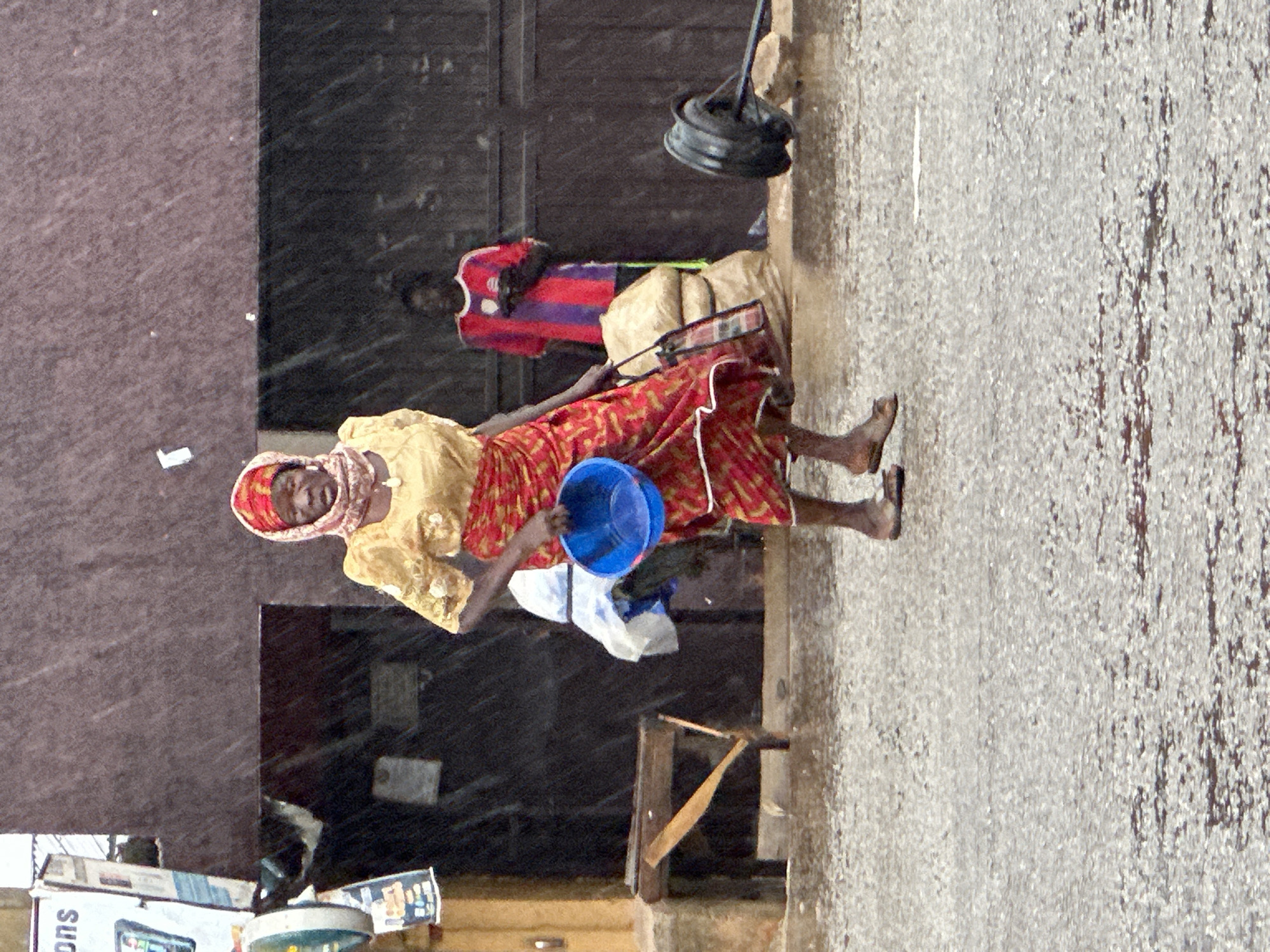 In Bukuru, Nigeria, during a downpour, a woman in a yellow top and a red and yellow patterned skirt walks across a wet asphalt street. She wears a matching headscarf and carries a blue bowl in her right hand and a shopping bag over her left shoulder. Behind her, a person in a red and purple striped shirt stands near a building with a dark facade. The ground is covered in puddles, and raindrops are visible in the air, indicating heavy rain. A wooden table and chair are positioned to the left, and a large tire lies on its side to the right.
