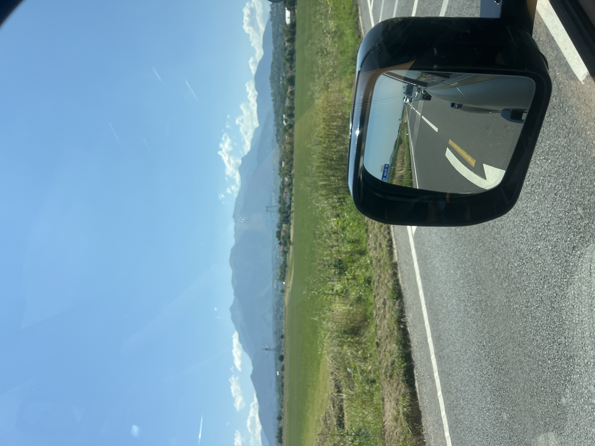 The image displays a scenic view captured from a moving vehicle. In the foreground, a car's side mirror reflects a highway with other vehicles. The highway stretches into the distance, bordered by a grassy verge and a field of green crops. In the background, a range of majestic mountains forms the horizon under a clear blue sky with scattered white clouds and faint contrails from aircraft. The scene appears to be in a rural or semi-rural area. The overall impression is one of travel on a bright, sunny day.