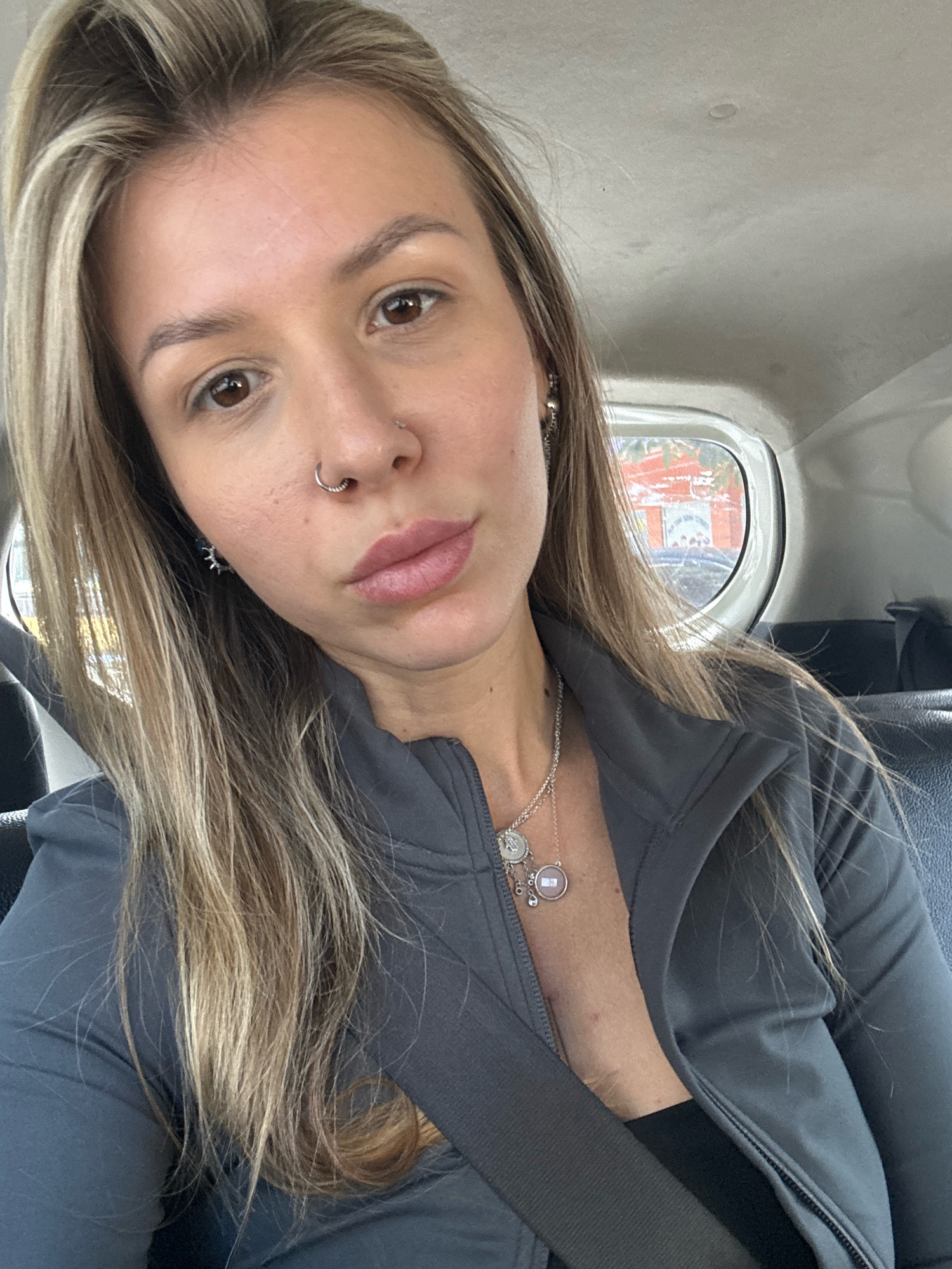 A woman with blonde hair and brown eyes is looking at the camera from inside a vehicle. She is wearing a gray zip-up jacket, a black top, and a necklace with multiple pendants. She also has a nose ring and earrings. The background shows the interior of the vehicle and a blurry view of buildings outside the window. The lighting suggests it is daytime.