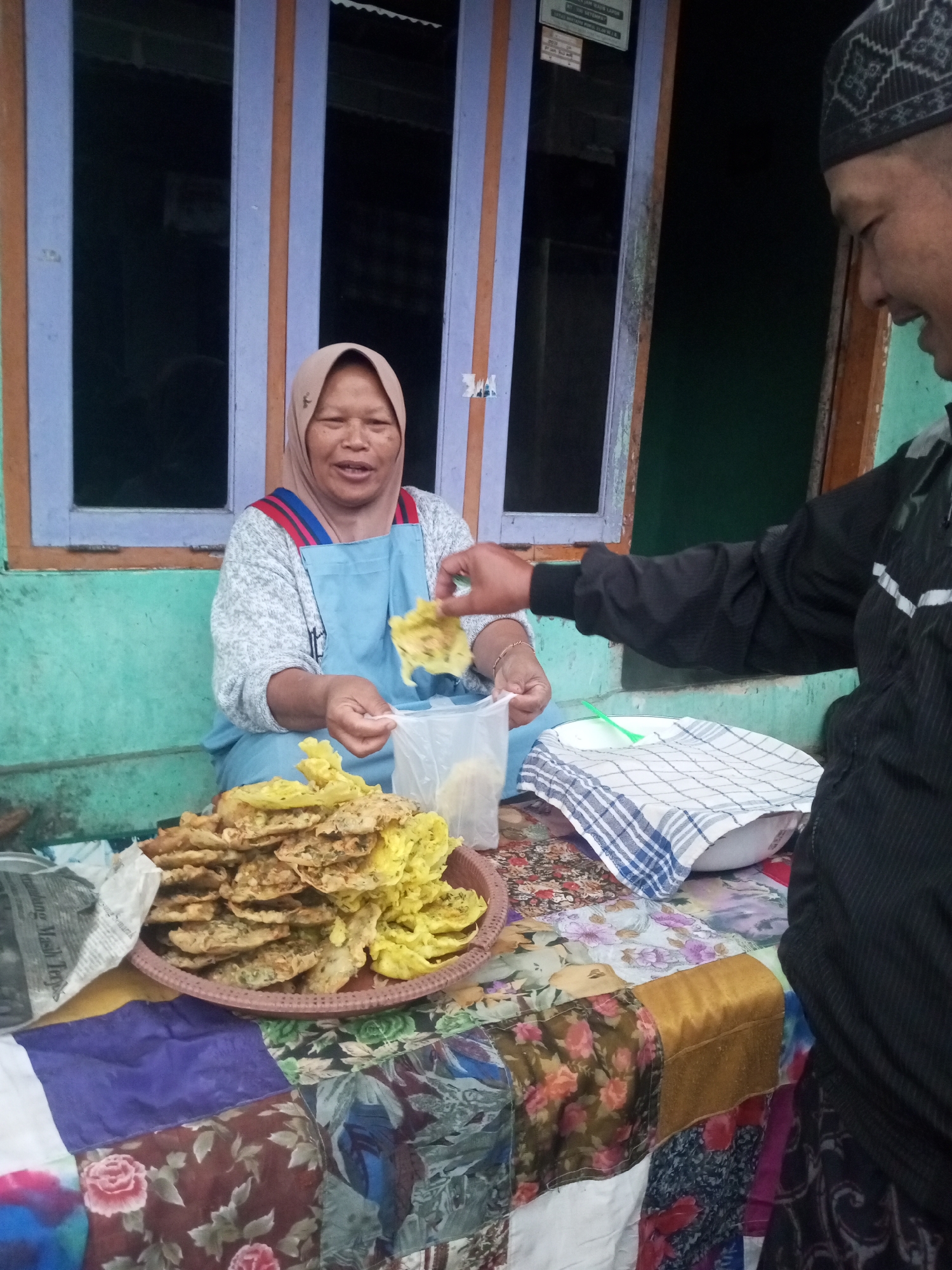 In Andongsili, Indonesia, a woman wearing a light brown hijab and a blue apron is seated, holding a fried food item. She has a light complexion and is smiling. In front of her, on a table covered with a colorful, patchwork cloth, is a brown woven basket filled with fried foods. Next to the basket is a clear plastic bag and a white bowl covered with a checkered cloth. To the right of the woman, a person wearing a black jacket and a patterned cap extends their right hand, also holding a fried food item, towards the woman. The background consists of a teal-colored wall with a window featuring purple and brown frames. The scene appears to be an outdoor food stall.