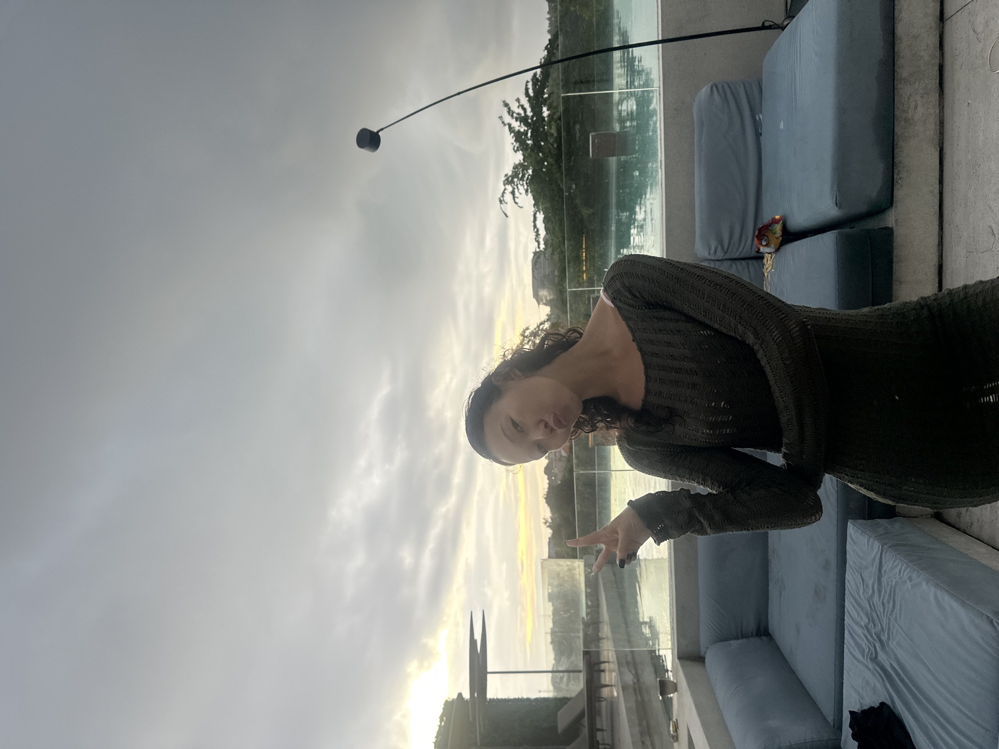 A woman is pictured outdoors on what appears to be a rooftop terrace. She is posing for the camera, making a peace sign with her right hand. She has curly dark hair and is wearing a dark, long-sleeved knit dress. The background features a cityscape partially obscured by cloudy skies and a body of water. The environment suggests a location in Kuta Selatan, Indonesia, as there is a visible structure that may be the hotel's infinity pool. There are light blue outdoor sofas on the terrace. A standing lamp with a curved neck is visible as well.