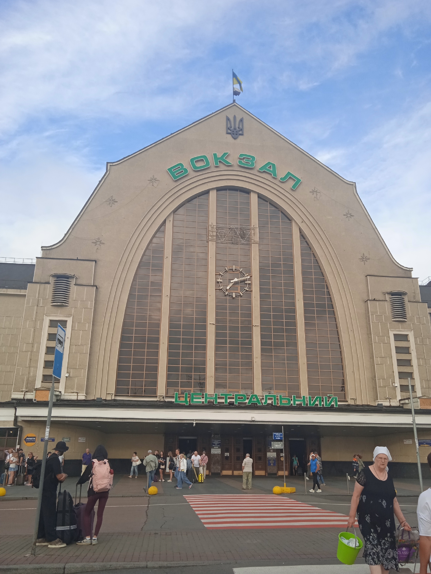 The exterior facade of a grand, arched train station building is shown under a partly cloudy sky. The building is a muted beige color with large, divided windows that reflect the sky. Green lettering across the top of the building reads "ВОКЗАЛ" which translates to "STATION". Below this, on the main entrance level, is more green lettering that reads "ЦЕНТРАЛЬНИЙ" which translates to "CENTRAL". A Ukrainian flag flies from a pole at the very apex of the building, and the Ukrainian trident emblem is visible just below the flag. A large clock face is visible on the central arch of the building. In front of the building, there is a pedestrian crossing with red and white stripes. People are seen walking across the street and gathered in front of the station entrances, some with luggage. A woman in a black dress and headscarf is crossing the street carrying a green bucket and shopping bags. Two people with luggage stand near a lamppost on the sidewalk. Several yellow bollards are placed around the pedestrian area.
