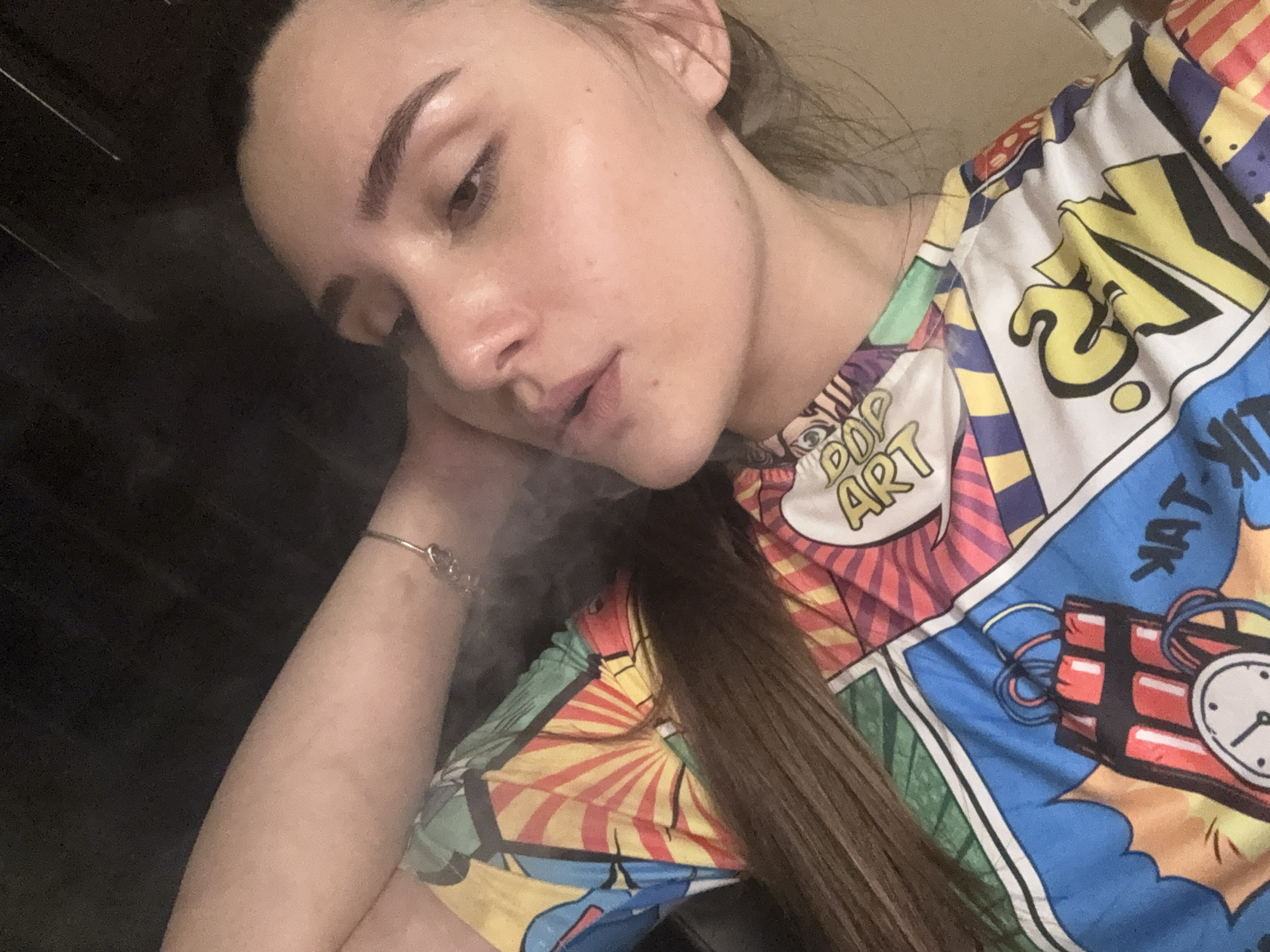 A close-up, high-angle shot captures a young woman lying down, her head resting on her left arm. She is wearing a brightly colored, comic-book-style t-shirt with a bold, graphic print featuring elements like a ticking bomb, the words "POP ART," and stylized letters. A thin, delicate bracelet adorns her left wrist. Wisps of smoke are visible around her mouth and nose, suggesting she may be exhaling. The lighting is dim, with the background appearing dark and out of focus. There is no discernible location in the city of Gabrovo, Bulgaria, or any other environmental context provided by the image.