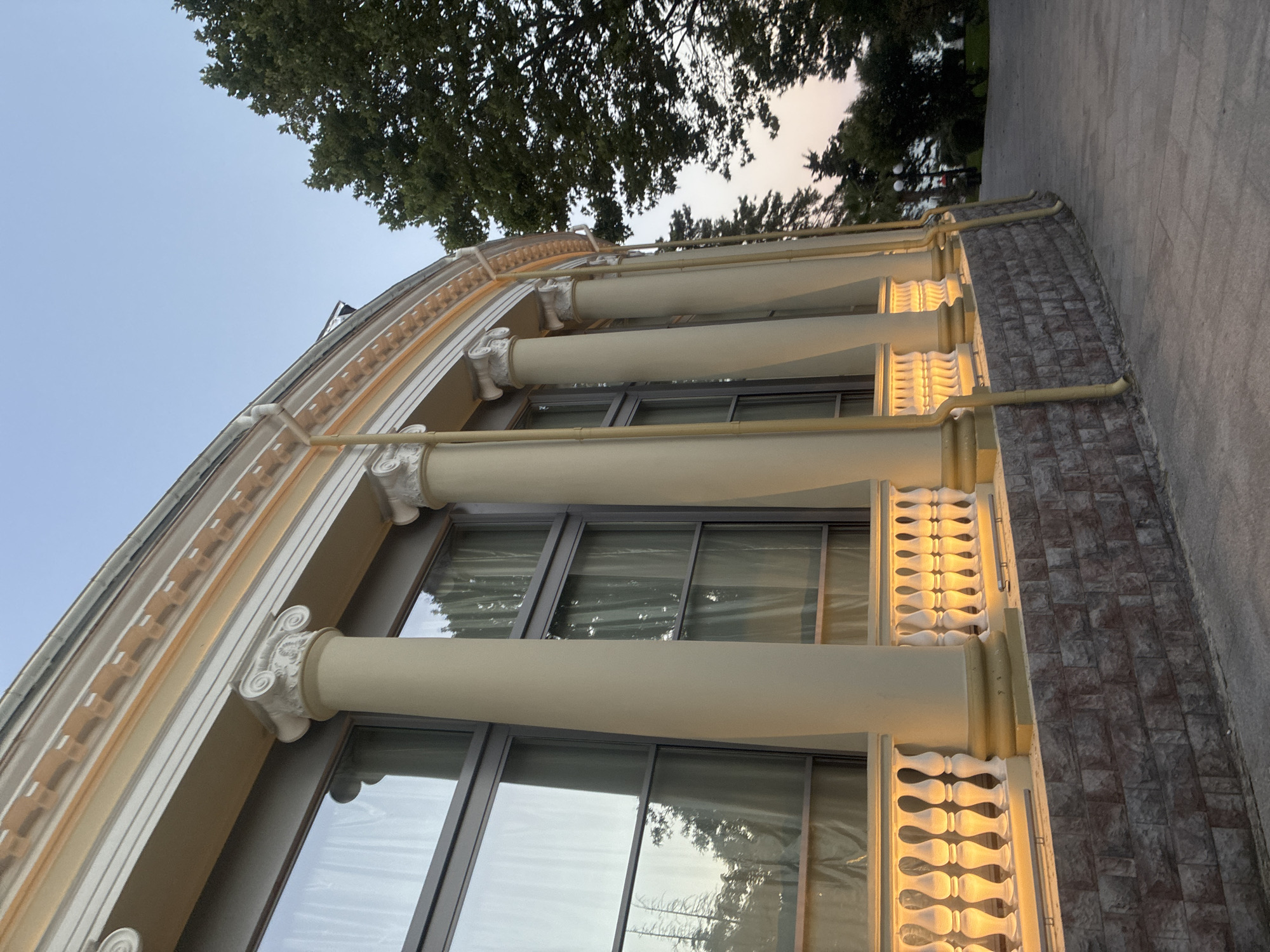 The image shows the exterior of a building with classical architectural elements in Sochi, Russia. The building features a curved facade with large windows and is supported by several large, cream-colored columns topped with Ionic capitals. A decorative white balustrade runs along the base of the windows, illuminated by warm lights from below. The lower part of the building is clad in stone blocks. To the right, lush green trees and a clear sky are visible, with hints of a park-like setting. The overall impression is of a well-maintained public building or establishment.