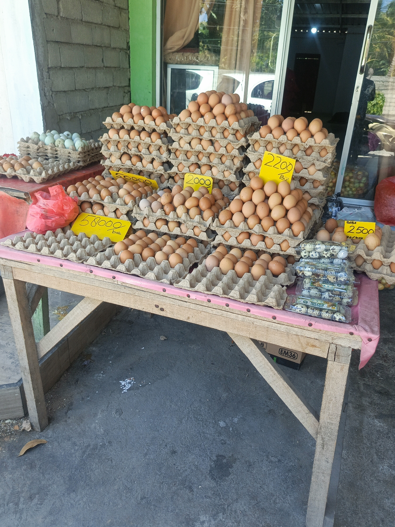 This image depicts an outdoor market stall in Tenggela, Indonesia, primarily featuring a variety of eggs for sale. The stall is set up on a wooden table draped with a pink cloth. Prominently displayed are numerous cartons of chicken eggs in various stages of stacking. Some cartons have yellow price tags with handwritten figures such as "58000", "2200", and "2500". To the left, a separate stack of lighter-colored eggs, possibly duck or quail eggs, is visible. In the center, a red plastic bag is placed on the table. To the right, several clear plastic containers filled with small, speckled eggs, likely quail eggs, are stacked. Behind the stall, a building with large glass windows is visible, reflecting the environment and showing glimpses of interior activity. The ground is paved with concrete, with some debris and a dry leaf scattered around the stall. The overall scene suggests a typical local market environment.