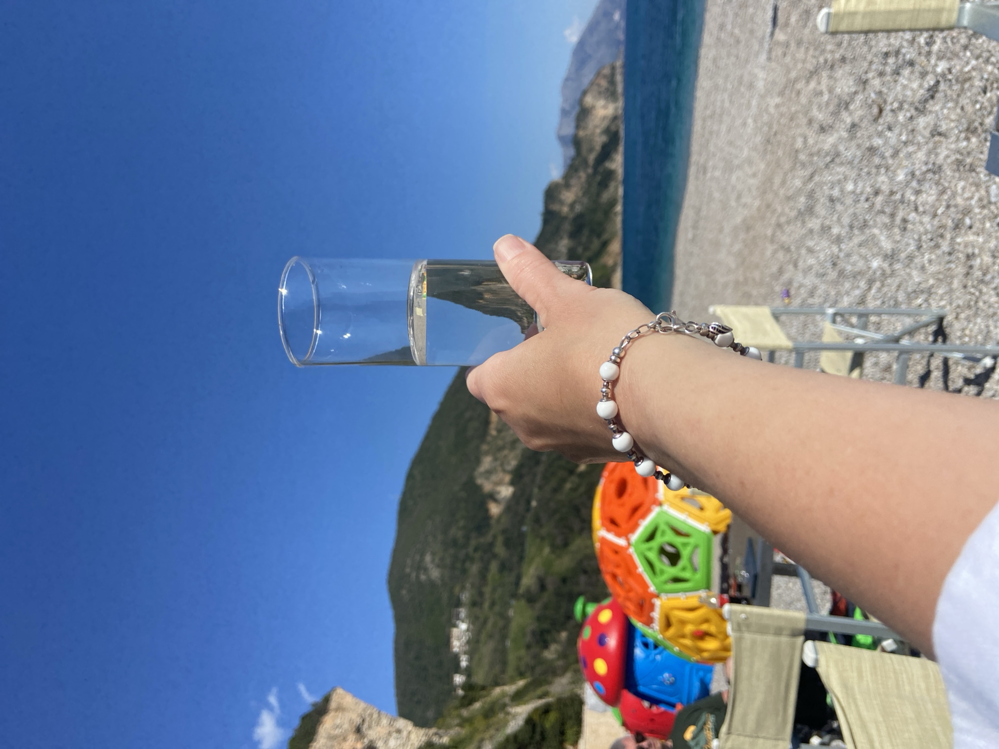 A person's arm holds a glass of liquid in front of a beach scene, with mountains and clear blue sky visible in the background. The location is Prijevor, Montenegro. The beach is pebbled, and there are beach chairs visible. The person's arm has a bracelet, and a colorful child's toy sits nearby.
