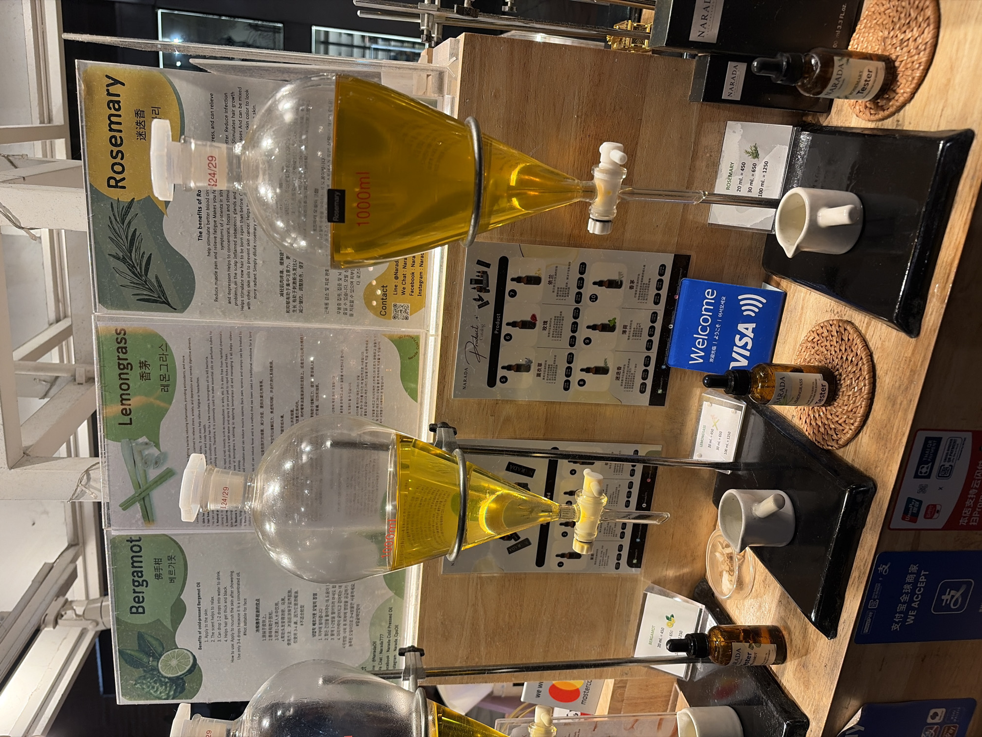 The image shows a display of essential oils in what appears to be a retail setting. Three large glass dispensers, each filled with a yellow oil, are prominently featured. Each dispenser is labeled with the name of an essential oil: Bergamot, Lemongrass, and Rosemary. Signs above the dispensers provide information about the benefits of each oil, with text in English and potentially other languages. Below the dispensers are small bottles of the oils, some labeled as "Tester," along with small white pitchers. The display is on a wooden counter, and there are payment signage for various methods like WeChat Pay, UnionPay, and Thai QR Payment visible. The context suggests this is a shop in Chiang Mai City Municipality, Thailand, likely selling natural or aromatherapy products. There are no people clearly visible in the immediate foreground. The overall impression is one of a product demonstration or retail display designed to attract customers.
