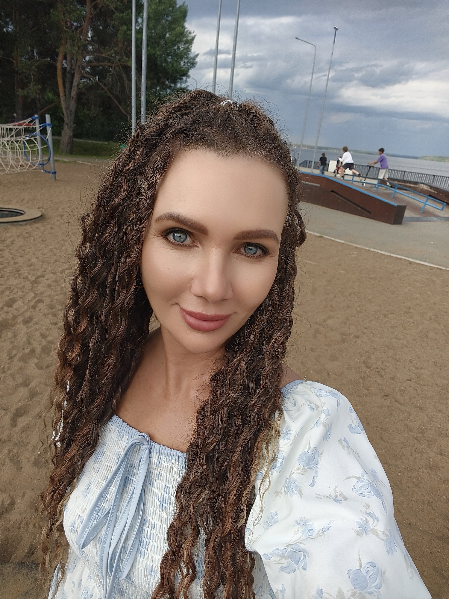 A selfie shows a woman with long, curly brown hair and bright blue eyes, smiling. She is wearing a white and blue floral dress. The background features a sandy area, a playground with a net structure, and a skate park with ramps. In the distance, several people are visible at the skate park, and a body of water can be seen. Tall poles with lights are also present. The sky is cloudy, suggesting an overcast or impending rain. The setting appears to be a recreational area near a waterfront. The overall mood is relaxed and pleasant. No text is visible.