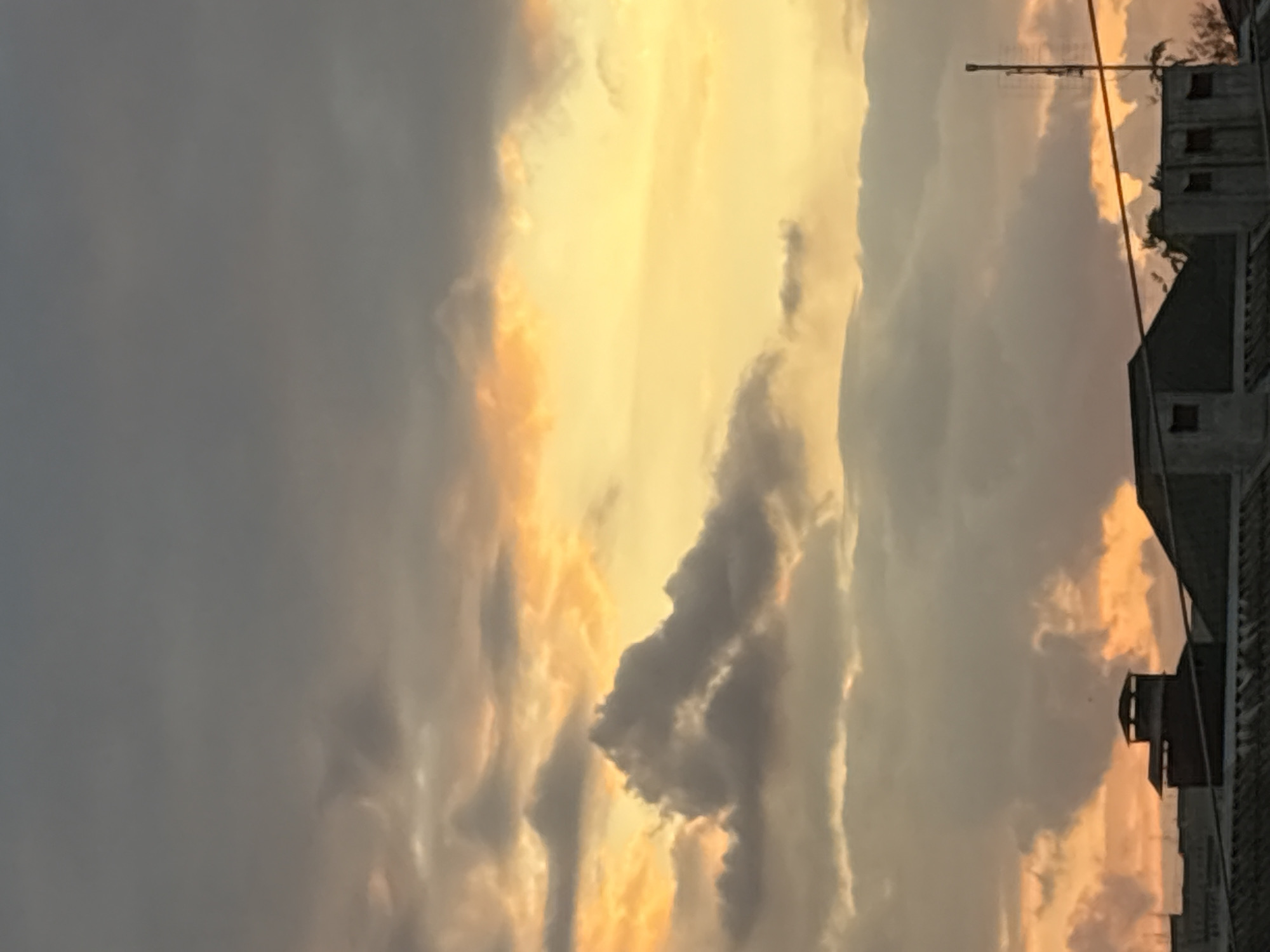 This is a photograph of the sky over the city of Georgiyevsk, Russia. The image captures a sunset with dramatic cloud formations. The sky is filled with a mix of dark gray and light gray clouds, interspersed with bright orange and yellow hues indicative of the setting sun. The lower portion of the image shows the rooftops of several buildings, including structures with chimneys and what appears to be an antenna. The overall impression is of a cloudy evening sky.