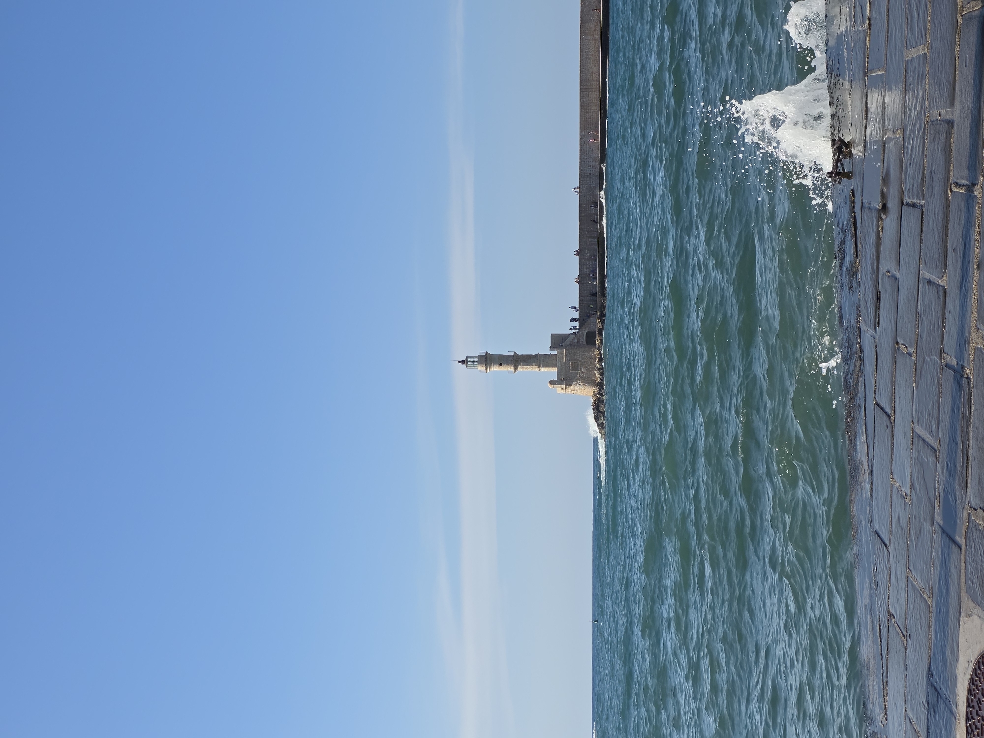 The image depicts the Chania Lighthouse, a prominent landmark located at the end of a breakwater in the Venetian harbor of Chania, Greece. The lighthouse, constructed of stone, stands tall against a clear blue sky. The rough sea, with waves crashing against the breakwater and creating white spray, suggests a dynamic coastal environment. Several people are visible walking along the breakwater, some near the lighthouse, indicating a public area. The foreground of the image is occupied by a stone-paved walkway, with a splash of water breaking on it. The overall scene captures the iconic view of the Chania Lighthouse and its surrounding harbor on a sunny day.