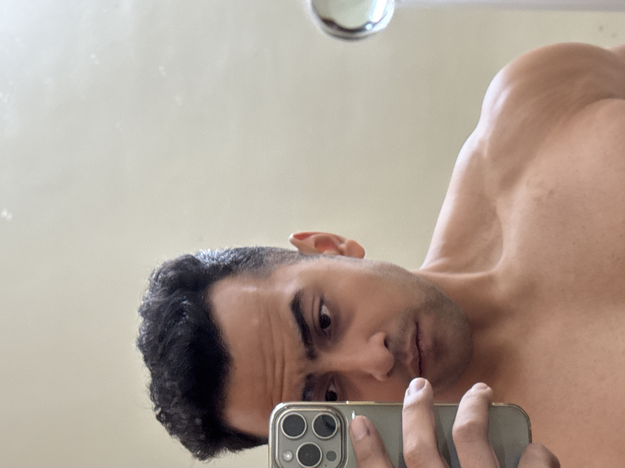 A shirtless man is taking a selfie in a bathroom mirror. He is holding a smartphone in his right hand, positioning it in front of his face. Part of a light fixture is visible near the top of the frame, above the man's right shoulder. The background is an off-white wall. The location is indeterminable from the image, so it's impossible to confirm it is in Kolkata, India.