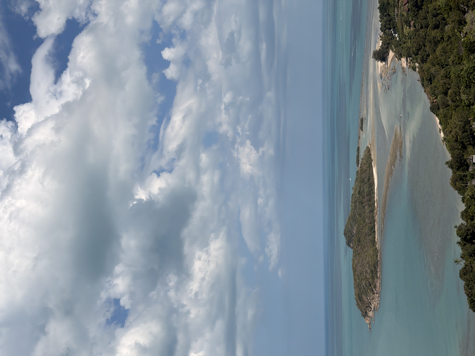 This is a high-angle, full shot of a tropical coastline in the Ko Samui District of Thailand. The sky is filled with a dramatic display of white and grey cumulus clouds, with patches of blue visible. The turquoise ocean stretches out to the horizon, meeting the hazy sky.
In the foreground, dense green foliage covers rolling hills. Closer to the water, there are scattered buildings, some appearing to be houses or resort accommodations, nestled amongst the trees. A sandy beach is visible along a curving bay.
Further out in the water, a large, treed island with rocky outcrops dominates the mid-ground. A sandbar or shallow reef connects this island to the mainland, creating a lagoon-like area. Numerous small fishing boats are moored near the shore on the right side of the frame. A single white sailboat is visible in the distance on the left. The overall scene conveys a serene and remote tropical landscape.