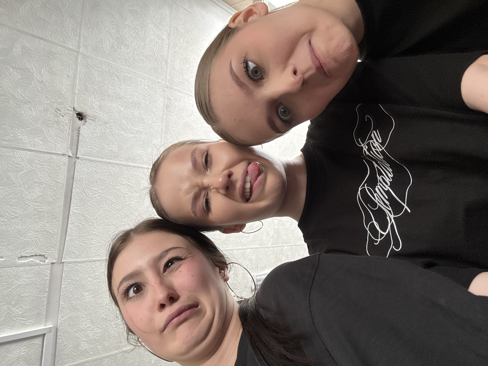 Three young women are posing closely together for a selfie-style photograph. They are positioned with their faces close to the camera, each making exaggerated facial expressions. The woman at the top of the frame has a wide-eyed expression and a slightly pursed mouth. The woman in the middle is sticking out her tongue and squinting. The woman at the bottom is looking upwards with a scrunched face. They are all wearing black shirts. The background appears to be a room with a textured ceiling and a light-colored wall. Based on the file name metadata, the location is Chaikovsky, Russia.