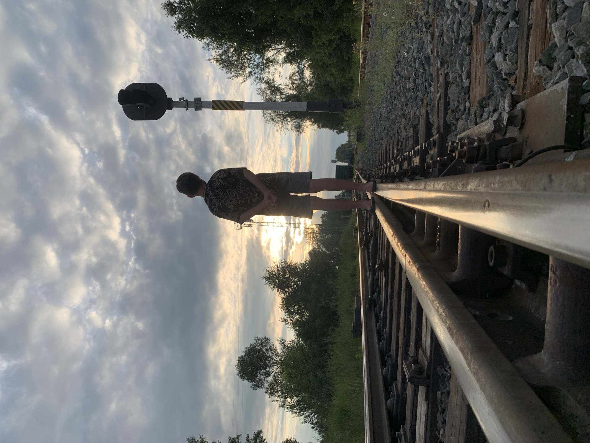 A young person stands on a railway track, facing away from the camera. They are wearing a patterned t-shirt and shorts. The railway track is lined with ballast and wooden sleepers. In the background, a railway signal stands tall with a black signal head and a striped pole. Trees and vegetation flank the tracks, with a cloudy sky overhead. The sun is setting, casting a warm glow through the clouds. The scene suggests a quiet, contemplative moment in a rural or semi-rural setting. The lighting and the presence of the railway infrastructure suggest it could be a disused or seldom-used track. The overall mood is serene and possibly a little melancholic. There is no visible text in the image. The location context is Chaikovsky, Russia.