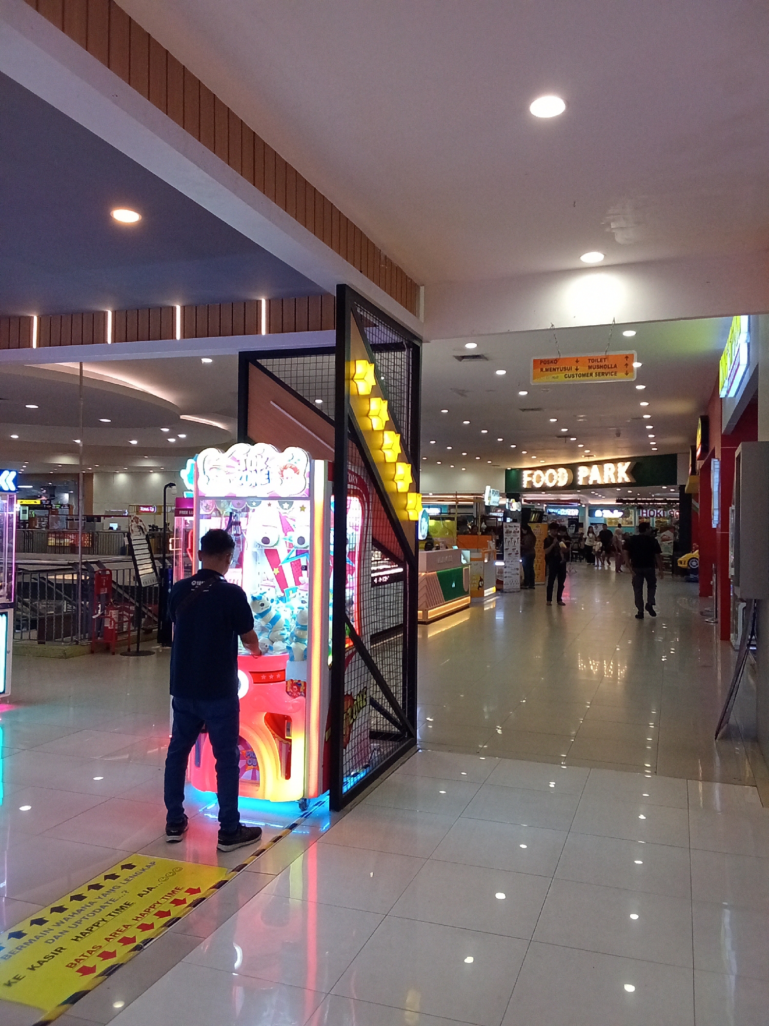 The image captures a section of an indoor public space, likely a shopping mall or entertainment center, in Kediri City, Indonesia. In the foreground, a man in a dark blue t-shirt and jeans stands in front of a brightly lit arcade machine, appearing to interact with it. The machine features cartoon characters and colorful lights. Yellow and black striped tape marks a boundary on the floor in front of the machine, with text in Indonesian that seems to indicate a designated area.
The environment is characterized by a tiled floor that reflects the ambient lighting, creating numerous bright spots. Above, the ceiling is a combination of flat white surfaces and a dark grey panel, with recessed circular lights illuminating the space. A large metal structure with wire mesh and yellow geometric accents is visible to the right of the arcade machine.
In the background, a walkway extends into the distance, lined with various shops and food establishments. A prominent green sign with "FOOD PARK" written in white letters is visible further down the walkway, indicating a food court area. Other signs, including one with directions to "POSKO", "TOILET", "R.MENYUSUI", "MUSHOLLA", and "CUSTOMER SERVICE," are also present, hanging from the ceiling. Several other individuals are seen walking in the background, some appearing to be patrons of the establishments. The overall impression is one of a well-lit, modern commercial area.
