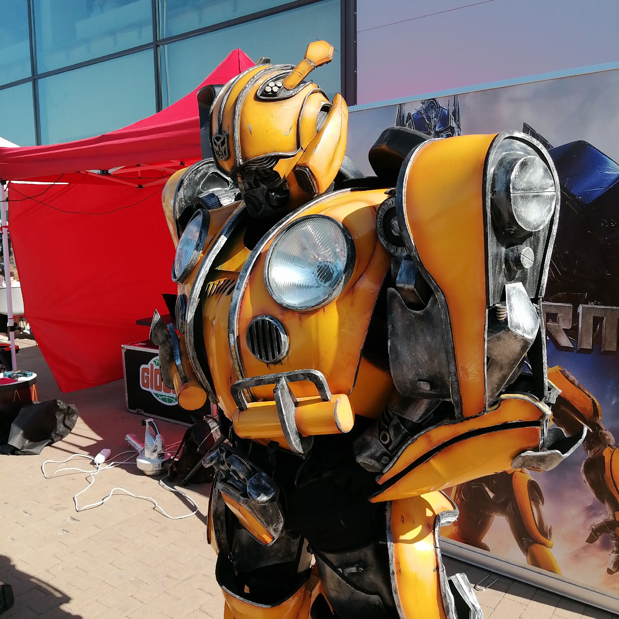 A detailed, full-body costume of Bumblebee from the Transformers franchise stands upright outdoors under bright sunlight in Yudino, Russia. The costume is primarily yellow with weathered dark gray and silver accents, mimicking battle damage and robotic parts. Its head features the iconic Autobot symbol on the left cheek, a dark eye visor, and an antenna-like protrusion on the top with small circular elements. The chest incorporates two large, ribbed circular headlights and a small rectangular grille-like vent below the left headlight. Visible black under-suit material shows between the segmented yellow and gray armor plates on the torso, arms, and legs.
In the background, a modern building with large, reflective blue-tinted glass windows is visible on the left. A bright red pop-up canopy or tent, supported by white poles, is partially seen behind the costume on the left side. To the right, a large promotional banner displays imagery from the Transformers franchise, featuring a blue and red robot (Optimus Prime) on the left part of the banner and a yellow-orange robot (likely Bumblebee) on the right, partially obscured by the costume's leg. Stylized text, partially visible, includes Cyrillic characters.
The ground is paved with light-colored rectangular concrete pavers. In the foreground and midground, various items are present: a black fabric bag, a small dark stand with minor objects, a black equipment case displaying a "Globus" logo, and a white power cord snaking across the pavers. The cord leads to a white electronic device, possibly a fan or fog machine, positioned near the base of the costume. Distinct shadows of the costume and other objects are cast on the paved ground, indicating direct sunlight from the upper right. No other individuals are visibly present beyond the person inside the costume.