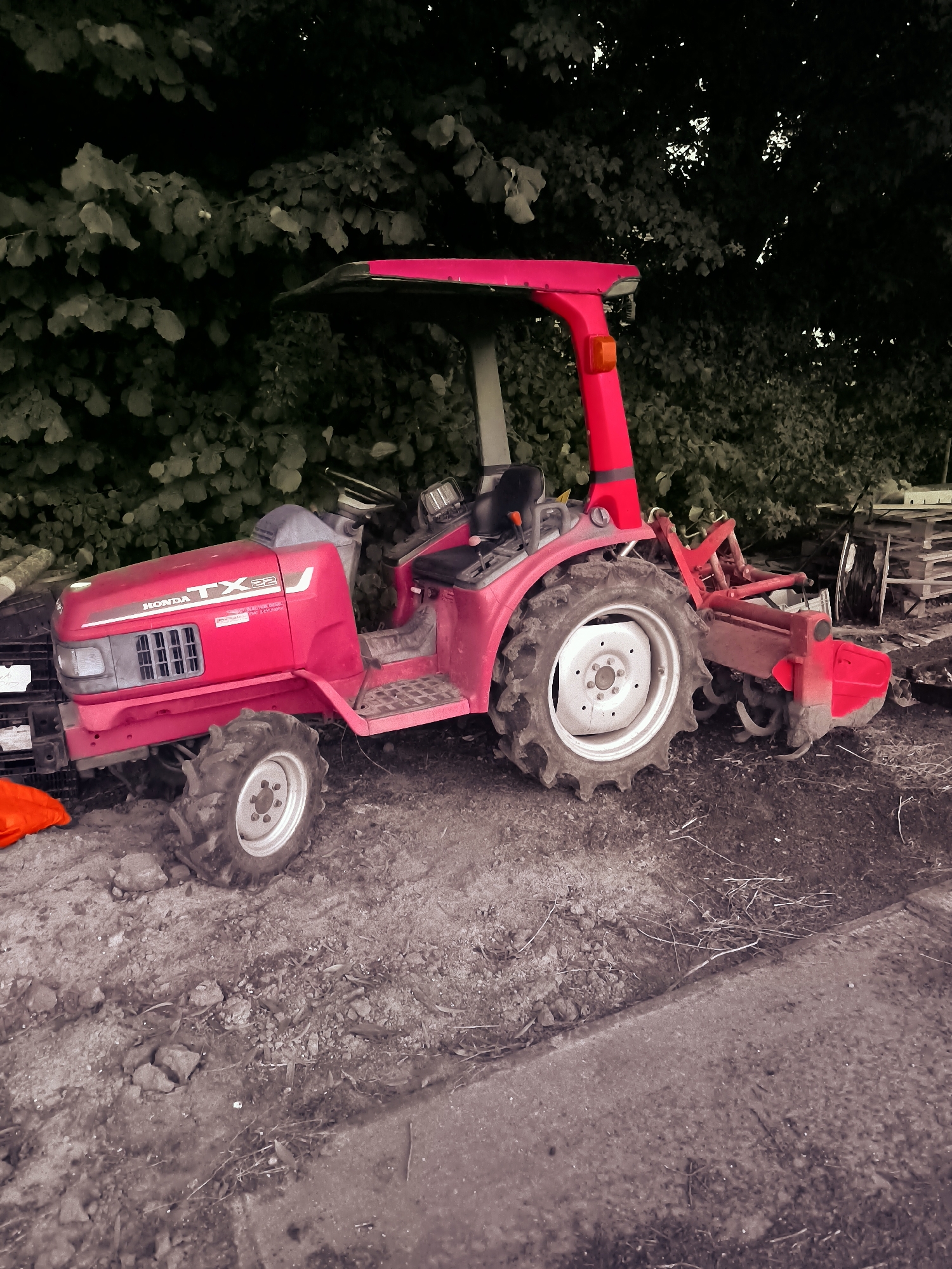 A bright red Honda TX22 tractor with a rotary tiller attachment is parked on a dirt and gravel surface beside a paved walkway, with lush green foliage forming a dark backdrop. The tractor has a driver's seat, steering wheel, and a red roll bar with a canopy. The front of the tractor features the "HONDA TX22" logo. To the left of the tractor, a bright orange object, possibly a cover or tarp, is visible. In the background, to the right of the tractor, stacks of wooden pallets are piled high. The scene appears to be outdoors, in a rural or semi-rural setting, possibly in Andijk, Netherlands, given the mention of the location. The ground is uneven and appears to be a mix of dirt, small stones, and dried leaves. The lighting suggests it is daytime, with some dappled sunlight filtering through the trees. There are no people visible in the image.