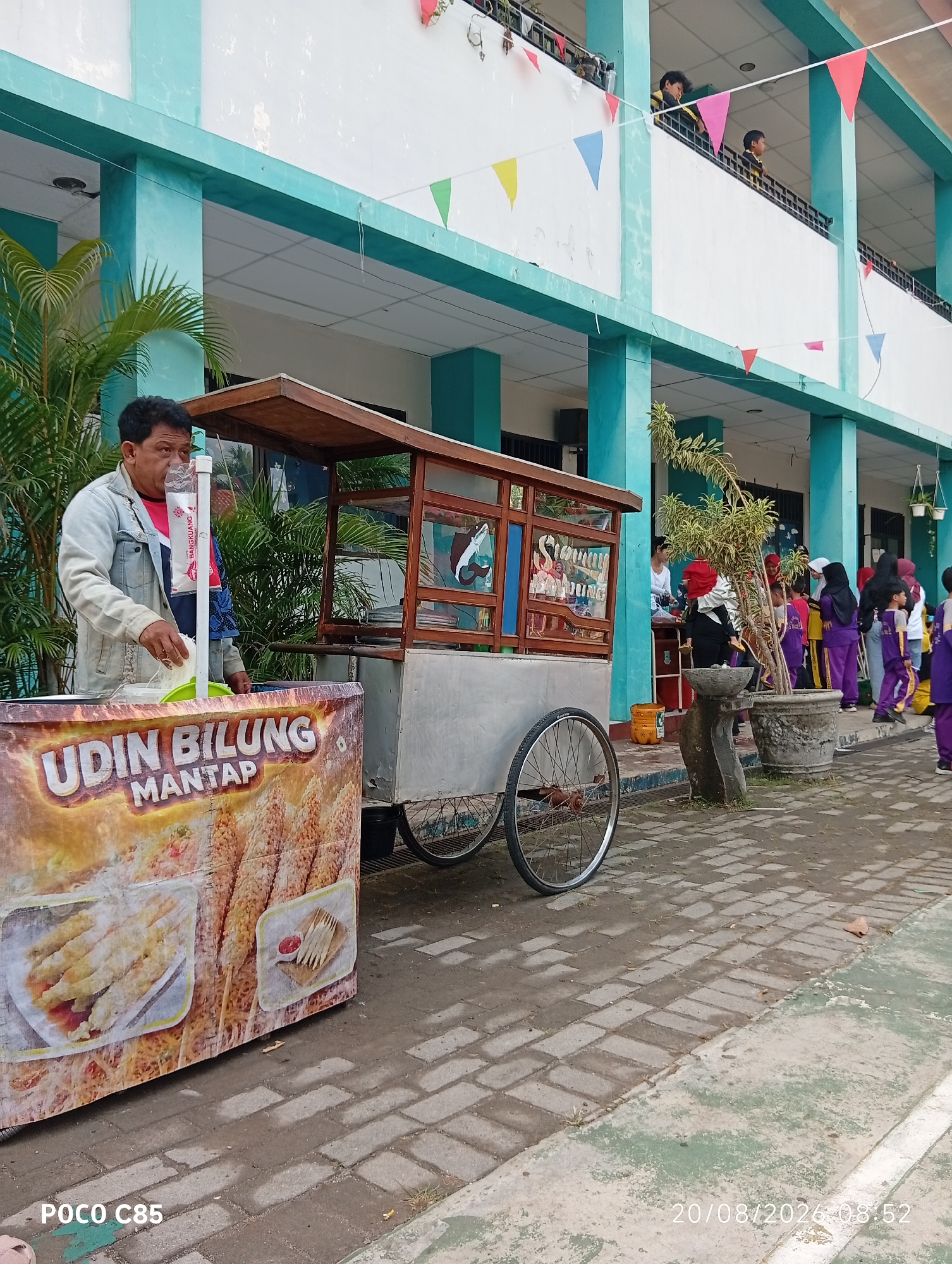 A man, wearing a light blue denim jacket over a red shirt, stands behind a food cart in an outdoor setting. The food cart has a sign that reads "UDIN BILUNG MANTAP" and displays images of fried food items. He is holding a white plastic bag, seemingly in the process of serving food. In the background, children in purple and yellow uniforms are seen standing near a building with turquoise accents and white walls, possibly a school. Colorful triangular flags are strung across the upper part of the building, suggesting a festive occasion. The ground is paved with interlocking bricks. The image includes a timestamp in the lower right corner: "20/08/2026 08:52". The presence of children in uniforms and flags suggests this is likely a school event or activity in South Tangerang, Indonesia.
