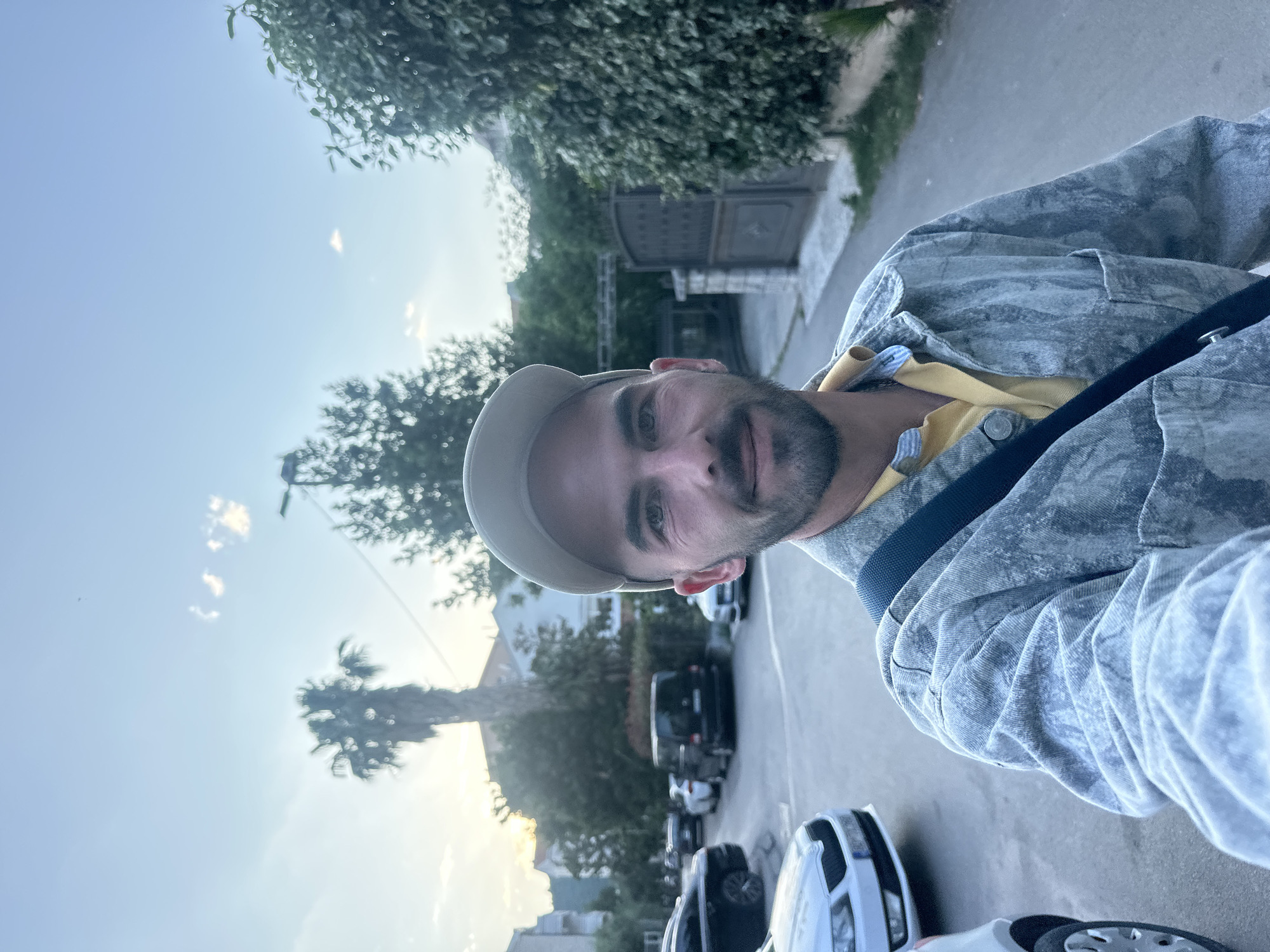 A selfie of a man is taken outside in Budva, Montenegro. He wears a tan baseball cap, a yellow polo shirt, and a light blue denim jacket with a black shoulder strap across his chest. The background shows a street with parked cars, a palm tree, and houses. In the far background, a stone wall and gate are visible. The sky is clear with a few clouds. The sun is setting.