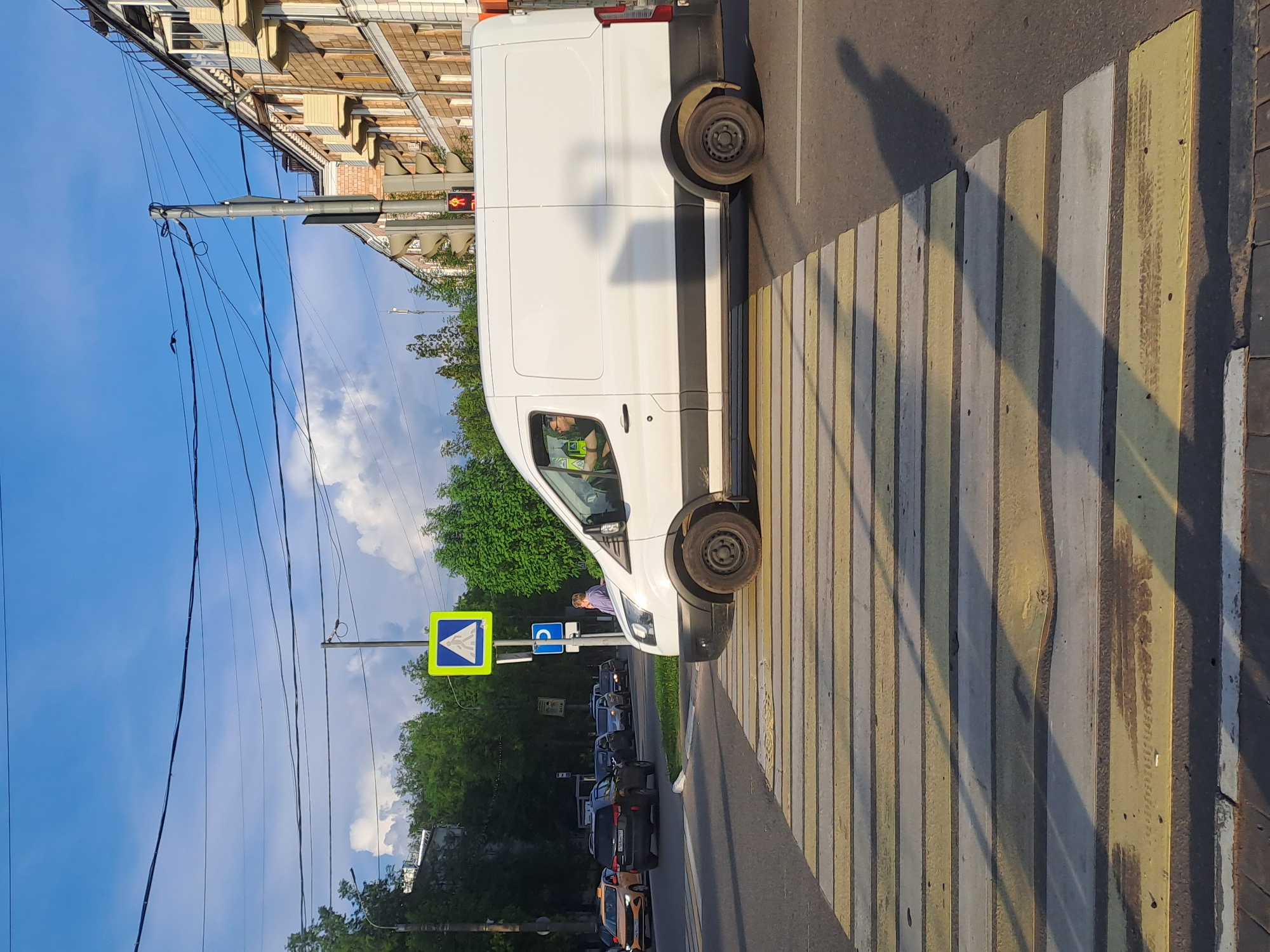 The image depicts a street scene in Elektrostal, Russia. A white van is stopped on a zebra crossing, partially blocking it. The crossing has alternating yellow and white stripes. A pedestrian crossing sign (blue square with a white figure walking on a zebra crossing) is visible near the van. In the background, a line of cars is waiting. Trees and a building are visible. A traffic light is showing red. Power lines run across the sky. The sky is blue with some clouds.