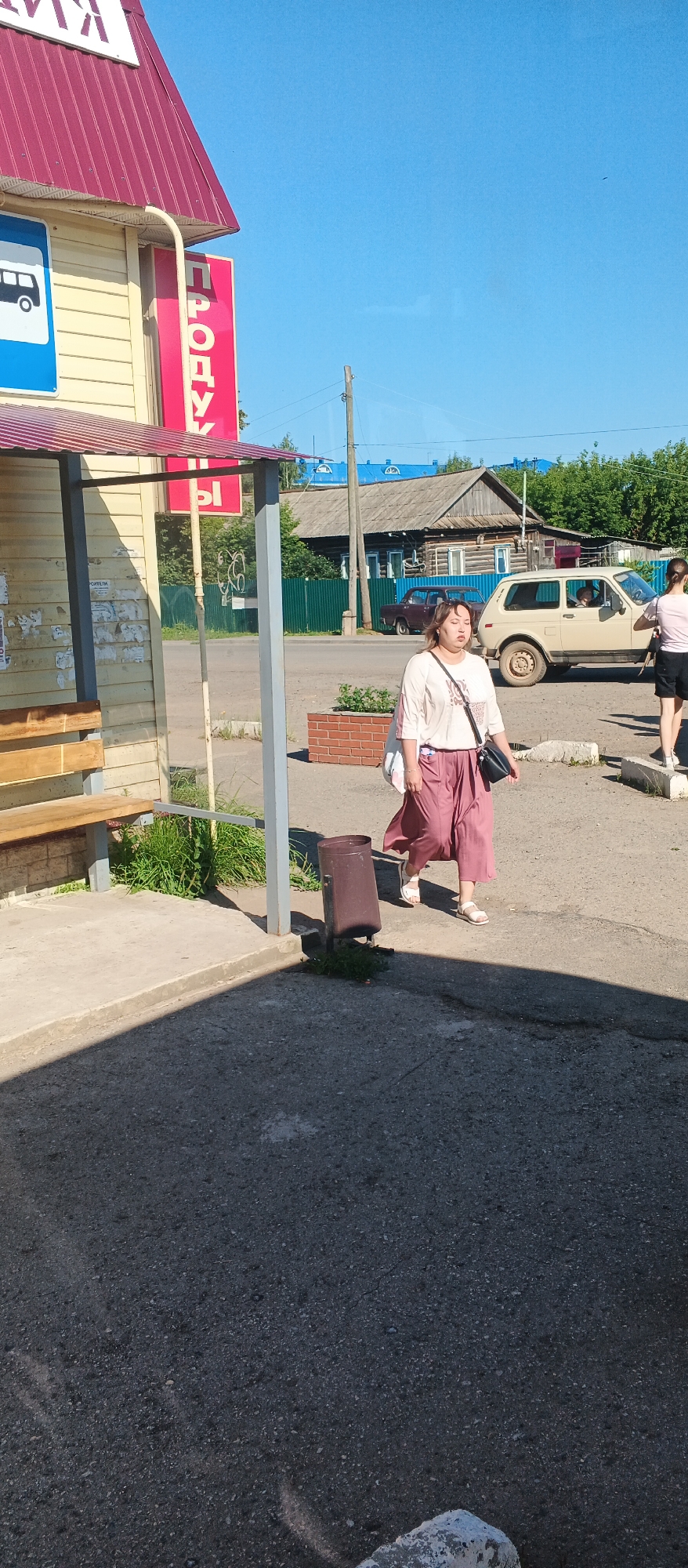 In the village of Kiyasovo, Russia, on a sunny day, a woman walks past a bus stop. The bus stop has a wooden bench and a red awning. A sign with the Cyrillic word "ПРОДУК" is visible on the side of the building. The woman, wearing a white blouse and a pink skirt, is carrying a black purse. Behind her, a beige car and a dark red car are parked on the street, with a wooden house and a green fence in the background. Another woman in a pink shirt and black shorts is walking away from the camera on the right side of the image. A trash can is located near the bus stop.