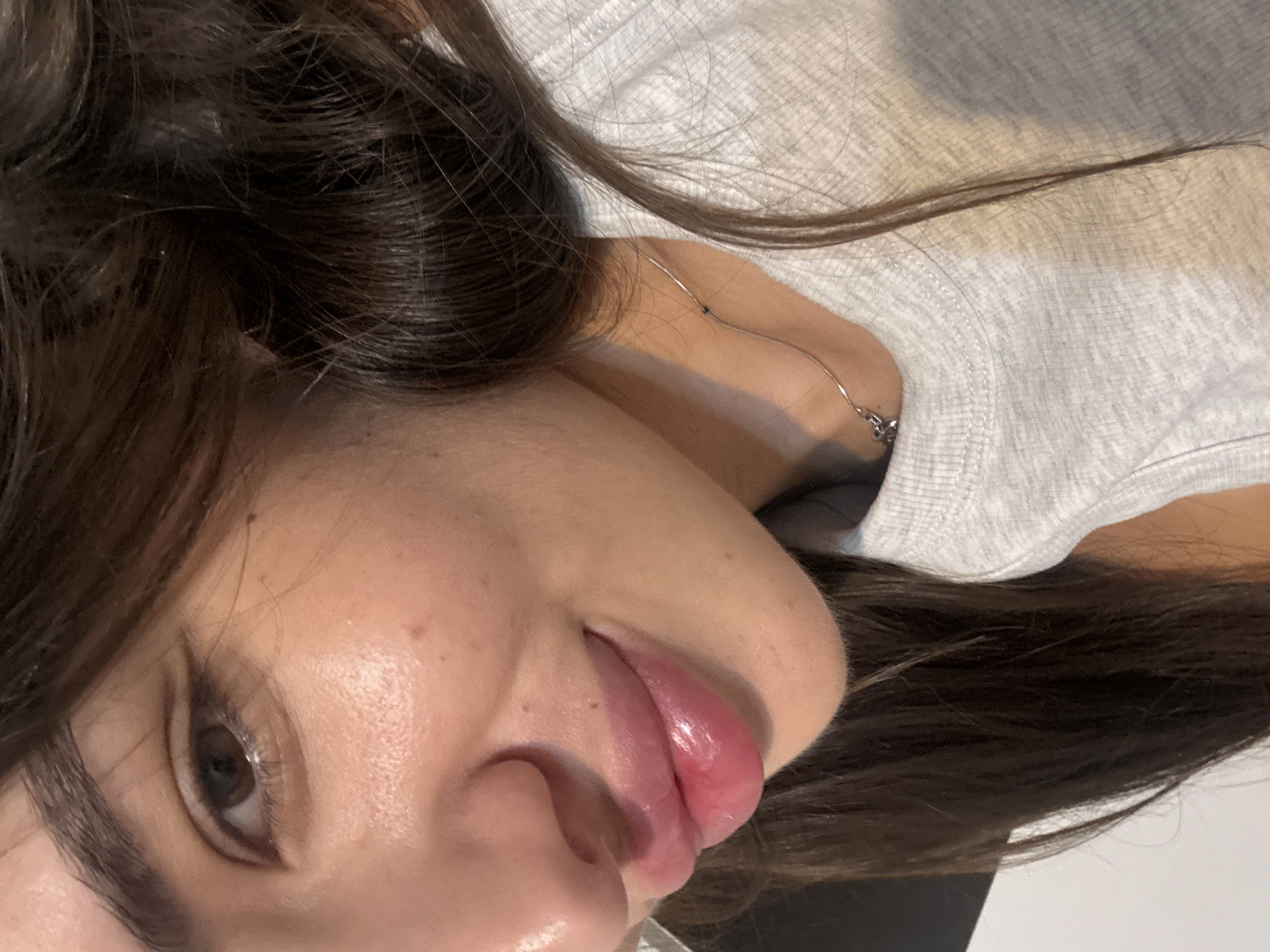 The image is a close-up, waist-up, portrait of a young woman. She has dark brown hair, brown eyes, and is wearing a light grey t-shirt. Her lips are plump and glossy, and she is wearing a thin silver necklace. The lighting suggests it is daytime, and the background is out of focus, appearing to be an indoor setting. There is no information available about the location of this image in the city of Chișinău, Moldova.