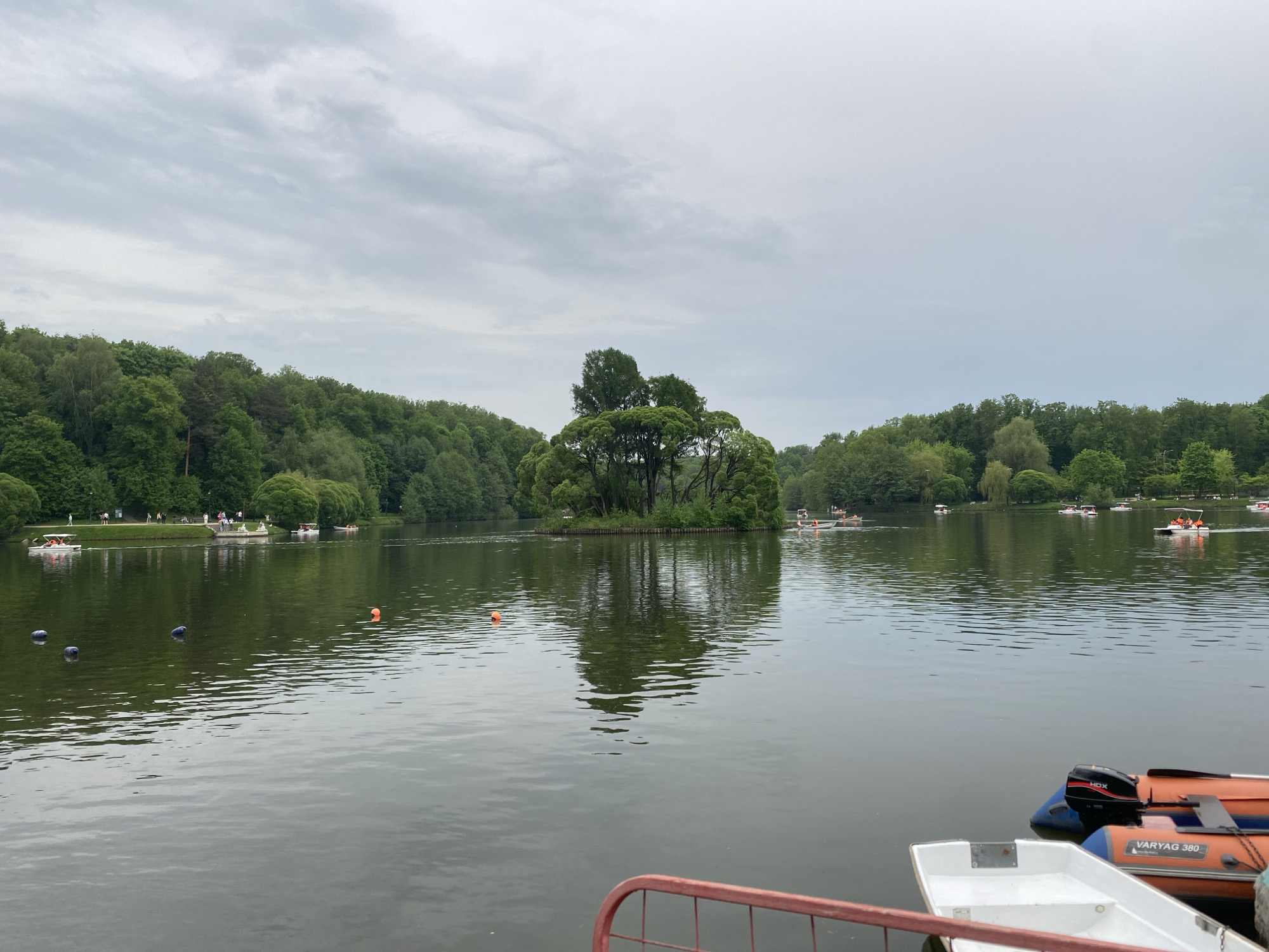 The image depicts a lake scene on a cloudy day. The location is in the city of Unknown, Unknown. In the center of the lake, there is a small island covered with trees. The reflection of the island and trees is visible in the water. Several small boats are scattered across the lake, with people visible in some of them. Orange buoys mark the area in the lake. The foreground shows portions of docked boats and a red railing. The banks of the lake are lined with dense green trees.