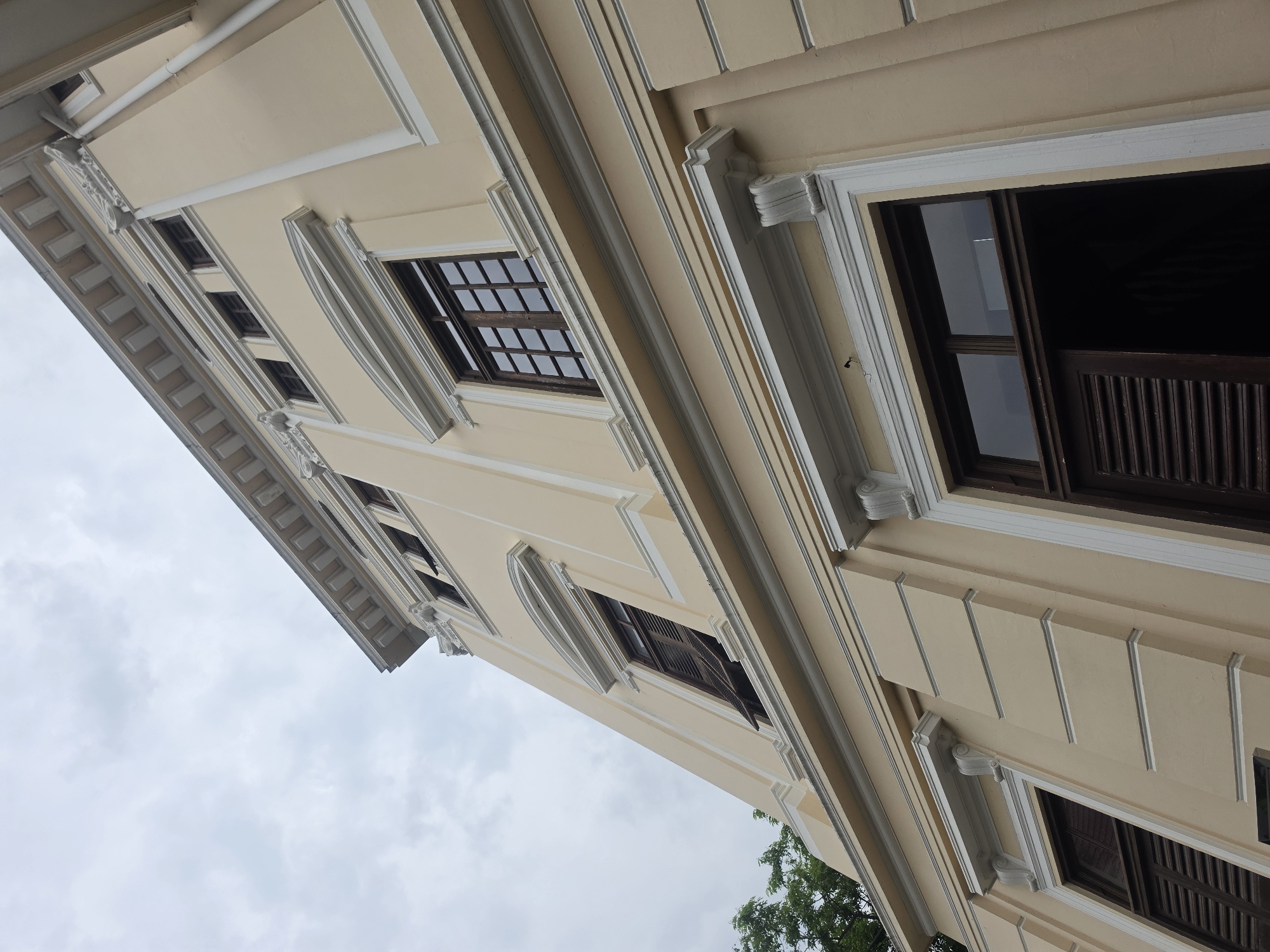 The image shows a low-angle view of a building in Bangkok, Thailand. The building is cream-colored with white architectural details, including decorative moldings around the windows and along the roofline. The windows are dark brown and appear to have multiple panes. Some windows are open, revealing dark interiors. The sky is overcast, and a portion of a tree is visible at the bottom of the image. The angle of the shot emphasizes the verticality of the building.