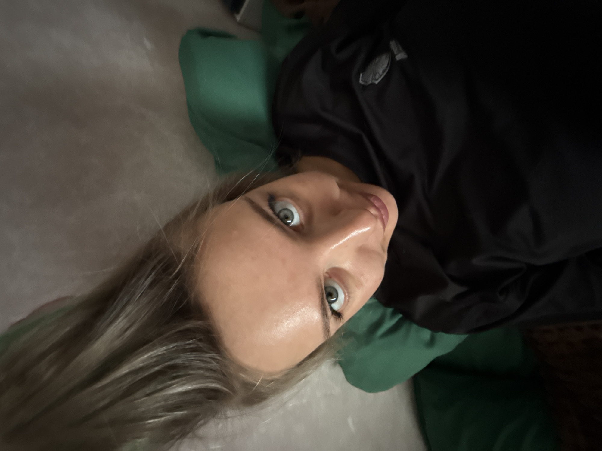 A woman with light blonde hair and blue eyes is lying on a grey-white surface, possibly a bed or couch. She is wearing a black jacket with a logo visible on the upper left chest area. The woman is positioned at an angle, with her head tilted towards the camera. She appears to be looking directly at the viewer with a surprised or slightly wide-eyed expression. She is lying on or partially supported by a green pillow or cushion.
There is no information available in the image to indicate the location is Borzya, Russia.